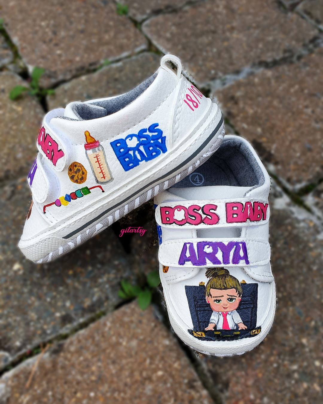 Custom hand-painted baby sneakers featuring Boss Baby theme. The shoes are white with colorful baby illustrations like pacifiers, bottles, cookies, and the name "Arya" painted in bold letters. Boss Baby characters are painted on the front, and birthdate details like "18/10/2020" and "October" are added on the back. Each pair is personalized, vibrant, and made for babies or toddlers as keepsakes or gifts.
