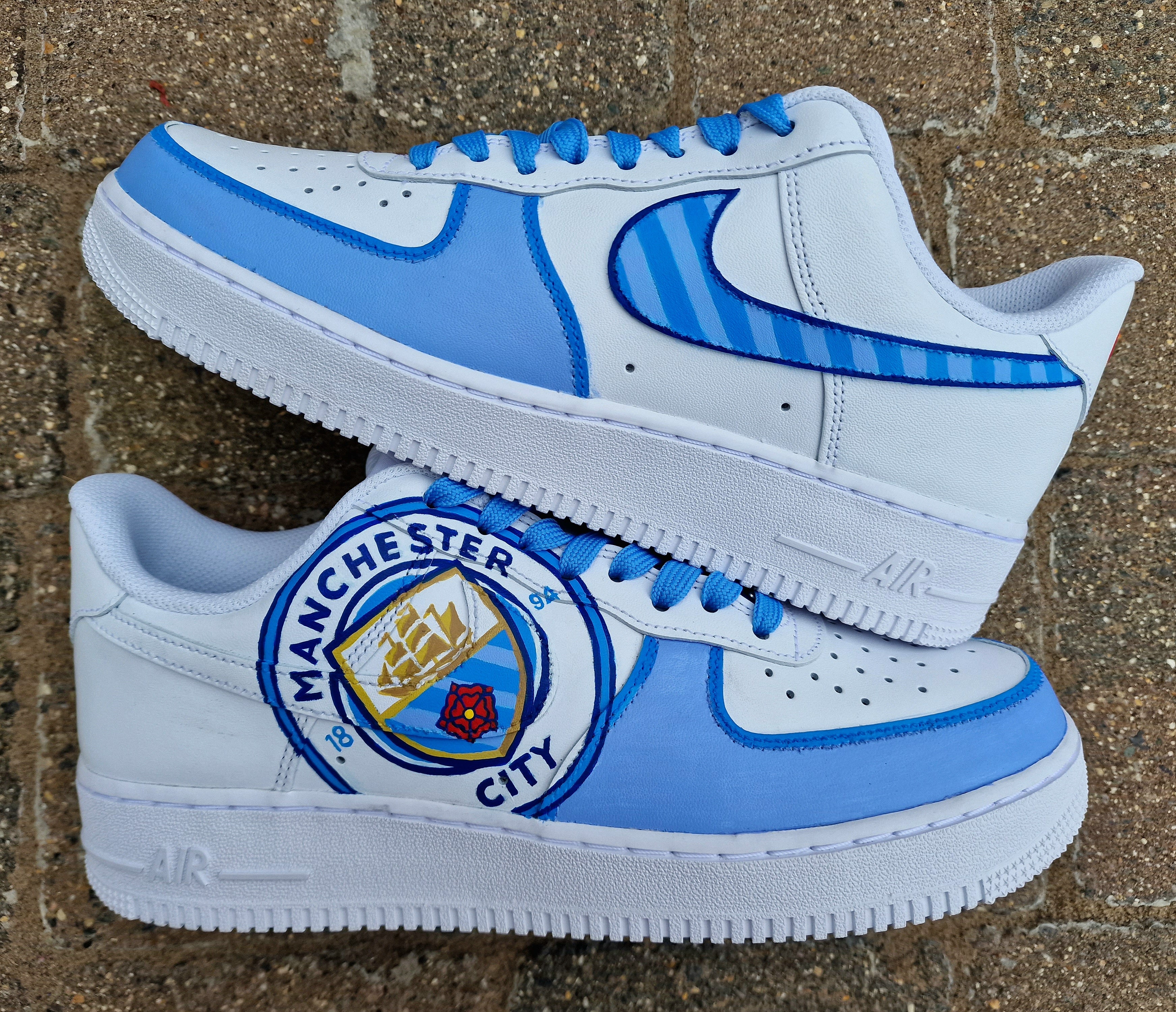 A pair of custom hand-painted Nike Air Force 1 sneakers featuring detailed artwork of the Manchester City Football Club crest, signature blue hues, striped swoosh, and personalized finishing, crafted by Gitartsy for football fans and sneaker collectors.