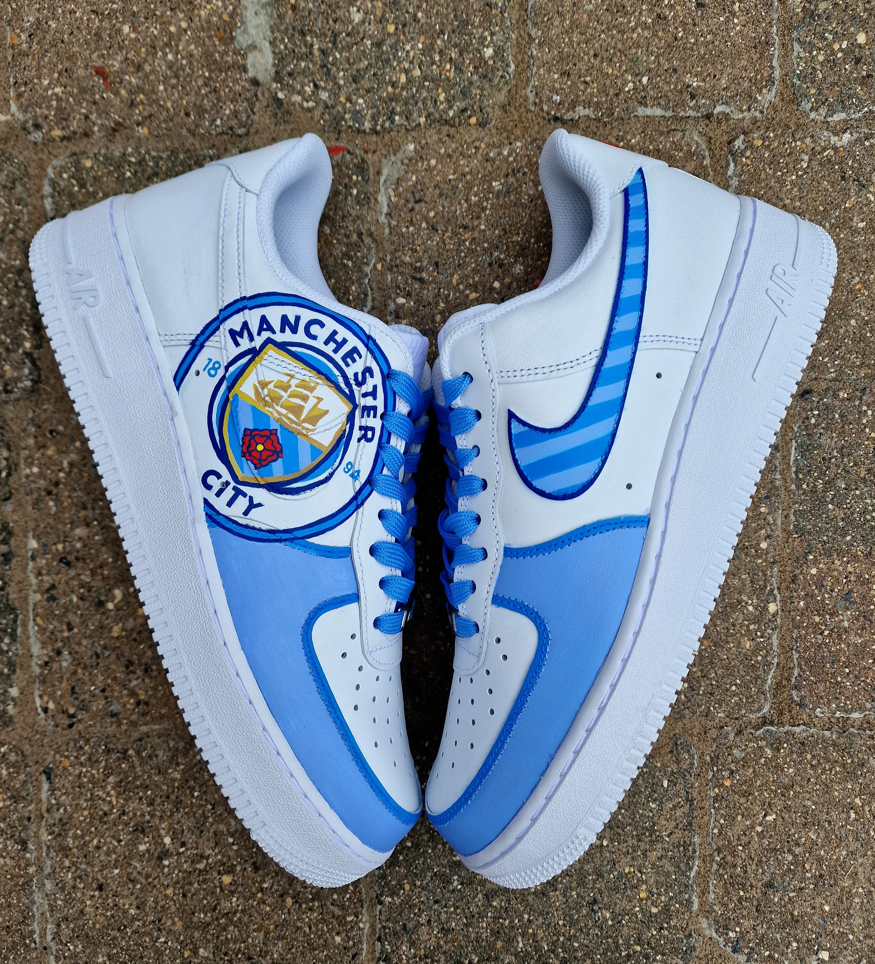 A pair of custom hand-painted Nike Air Force 1 sneakers featuring detailed artwork of the Manchester City Football Club crest, signature blue hues, striped swoosh, and personalized finishing, crafted by Gitartsy for football fans and sneaker collectors.
