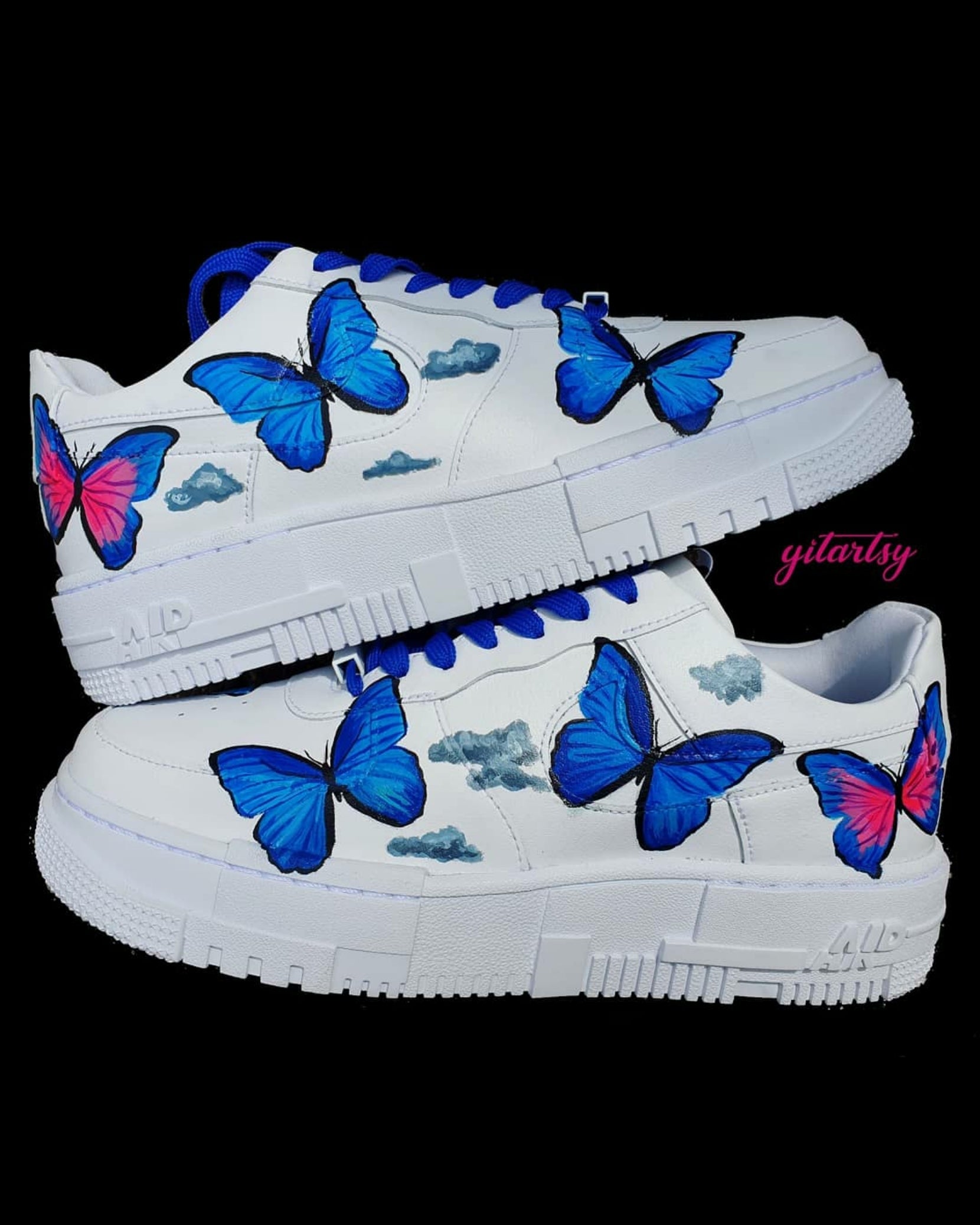 Hand-painted custom Nike Air Force 1 sneakers featuring vibrant blue and pink butterflies fluttering among soft grey clouds, set against a crisp white leather background. The design is detailed and symmetrical across both shoes, with bold black outlines emphasizing the wings. Finished with either bright pink or royal blue laces, the sneakers showcase a clean, street-style aesthetic with an artistic twist.