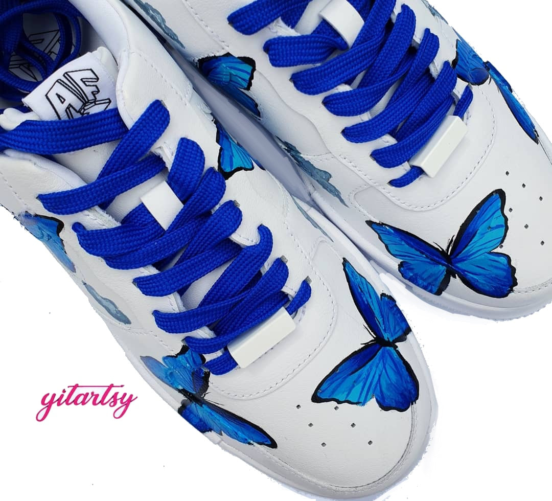 Hand-painted custom Nike Air Force 1 sneakers featuring vibrant blue and pink butterflies fluttering among soft grey clouds, set against a crisp white leather background. The design is detailed and symmetrical across both shoes, with bold black outlines emphasizing the wings. Finished with either bright pink or royal blue laces, the sneakers showcase a clean, street-style aesthetic with an artistic twist.
