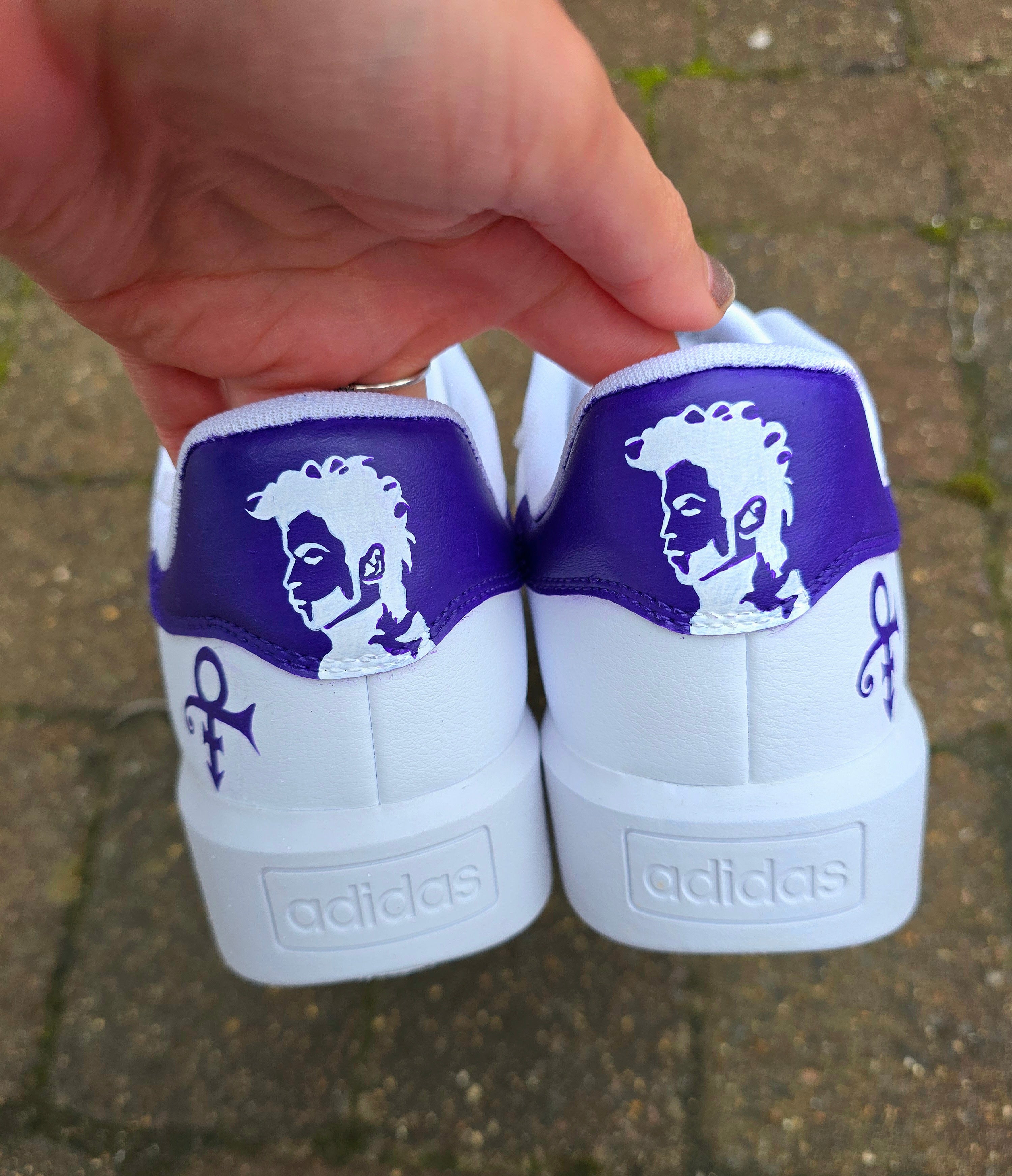 Custom hand-painted Adidas sneakers featuring a vivid purple tribute to the legendary singer Prince, including his iconic symbol, stylized signature, and portrait art on a clean white leather base with purple stripes.
