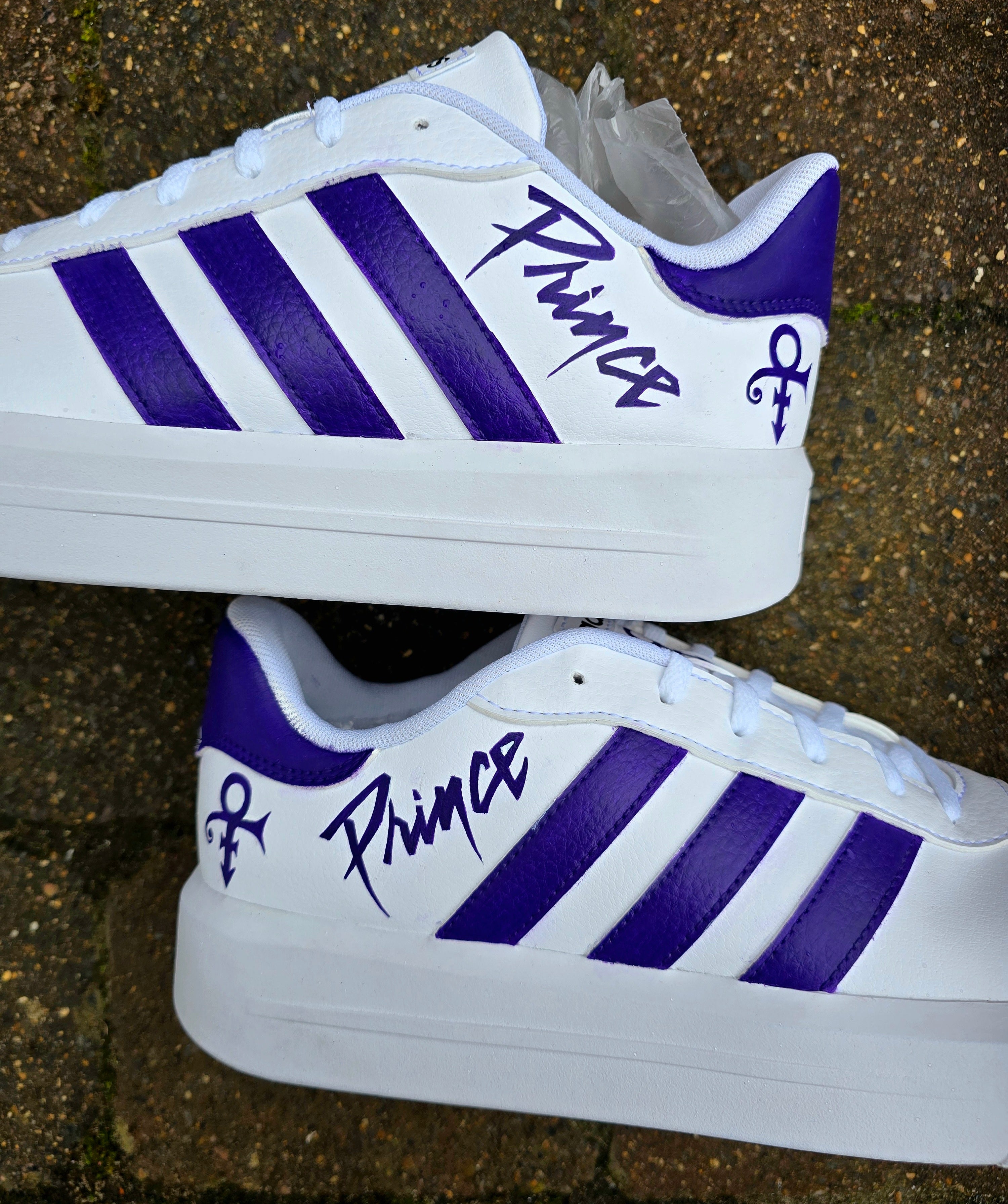 Custom hand-painted Adidas sneakers featuring a vivid purple tribute to the legendary singer Prince, including his iconic symbol, stylized signature, and portrait art on a clean white leather base with purple stripes.