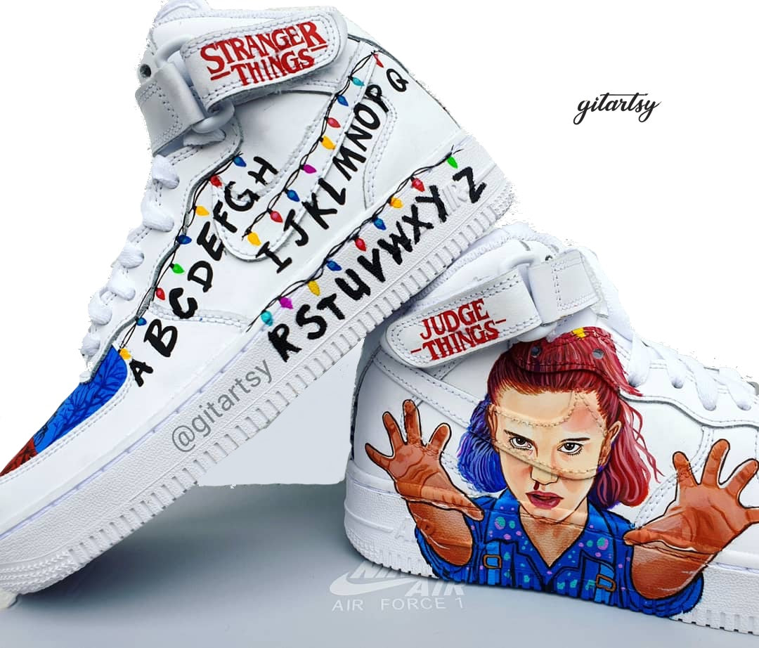 Hand-painted custom Nike Air Force 1 sneakers inspired by the Stranger Things Netflix series. The design features iconic elements including a realistic portrait of Eleven with her hands raised in a telekinetic pose, the alphabet wall with colorful string lights, red and blue toe panels depicting silhouette scenes of the kids on bikes, and high-top details like "Stranger Things" and "Judge Things" logos. Painted on a white leather Nike AF1 base with durable and vibrant colors.