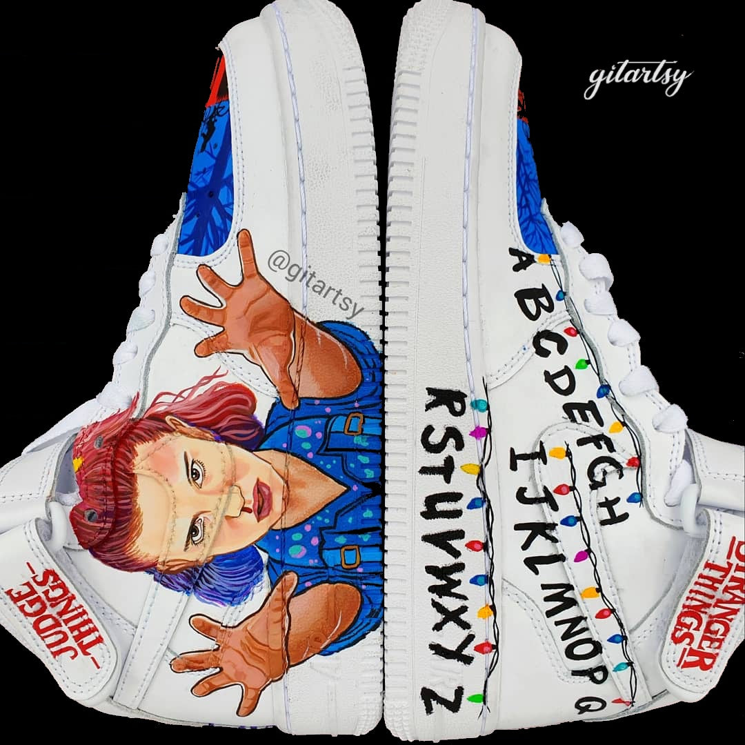 Hand-painted custom Nike Air Force 1 sneakers inspired by the Stranger Things Netflix series. The design features iconic elements including a realistic portrait of Eleven with her hands raised in a telekinetic pose, the alphabet wall with colorful string lights, red and blue toe panels depicting silhouette scenes of the kids on bikes, and high-top details like "Stranger Things" and "Judge Things" logos. Painted on a white leather Nike AF1 base with durable and vibrant colors.