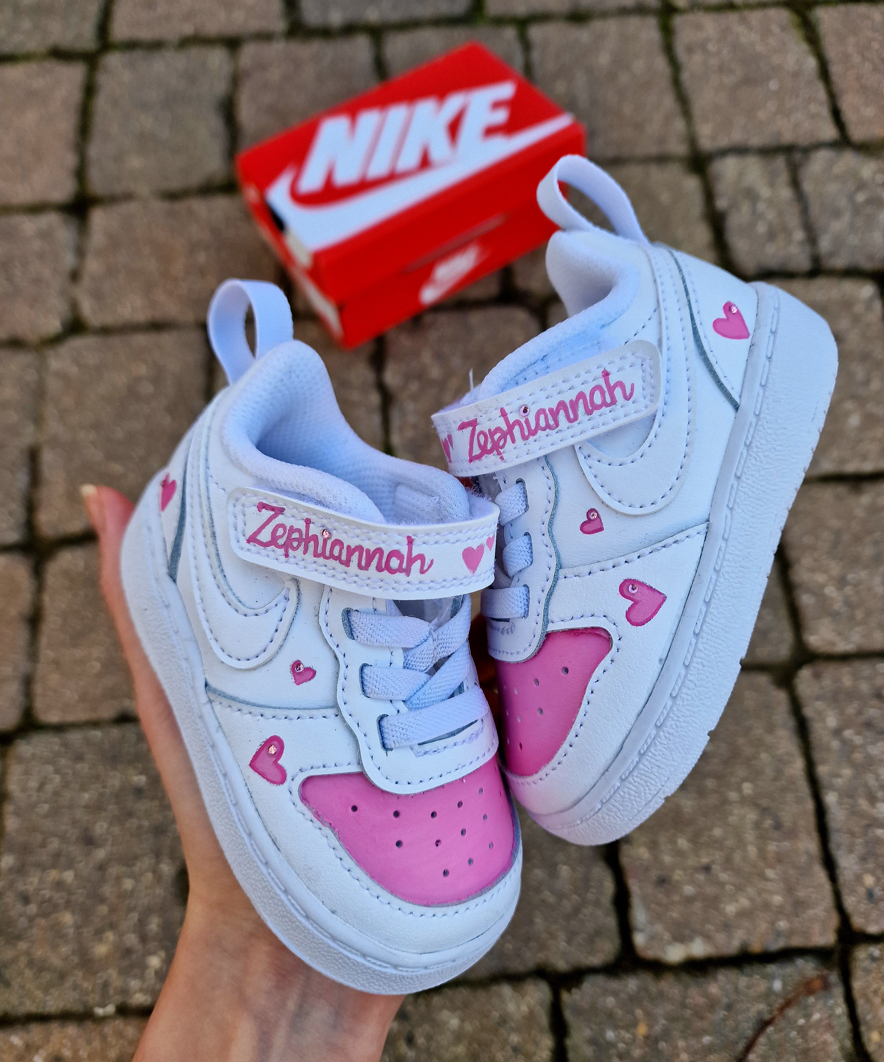 Custom Nike baby sneakers hand-painted in white and pink, featuring a personalized name "Zephaniah" on the strap and tongue. Accented with pink hearts on the side panels and heel. The sneakers sit on a red Nike shoe box on a stone pavement. Perfectly crafted for kids with durable materials and cute customization for special occasions.