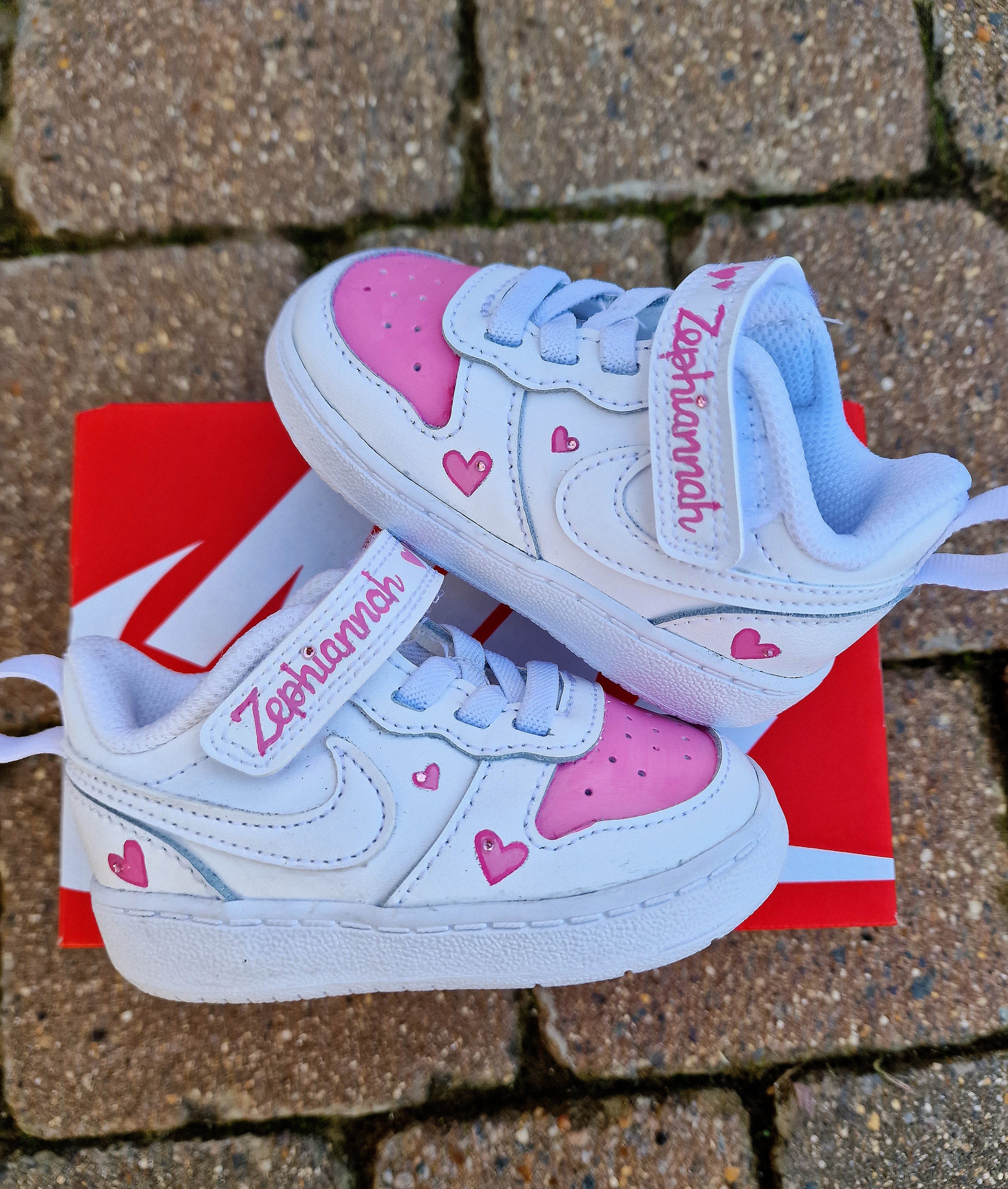 Custom Nike baby sneakers hand-painted in white and pink, featuring a personalized name "Zephaniah" on the strap and tongue. Accented with pink hearts on the side panels and heel. The sneakers sit on a red Nike shoe box on a stone pavement. Perfectly crafted for kids with durable materials and cute customization for special occasions.