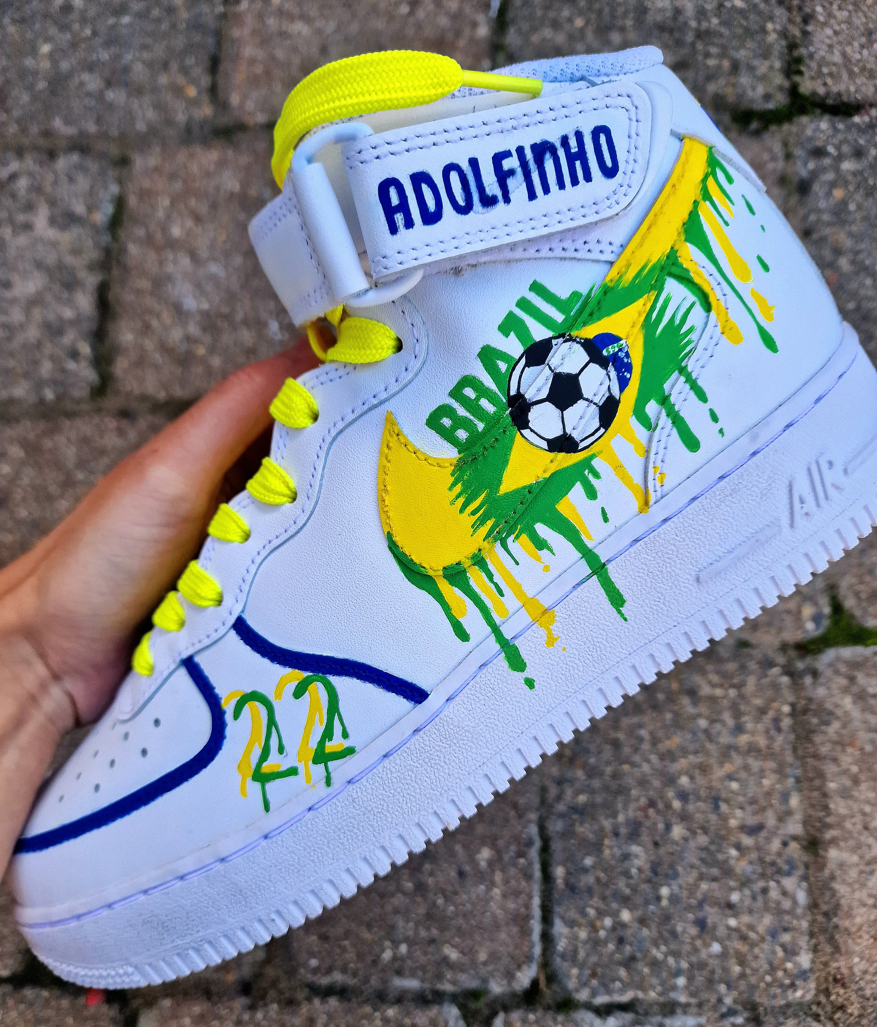 Custom Adolfinho Brazil-inspired Nike Air Force 1 sneakers featuring hand-painted green, yellow, and blue drip art, football graphics, the player name Adolfinho, the number 22, custom strap text, and vivid laces; unique wearable streetwear art designed for football fans and sneaker collectors who want personalised, one-of-a-kind kicks that celebrate Brazilian sports culture.