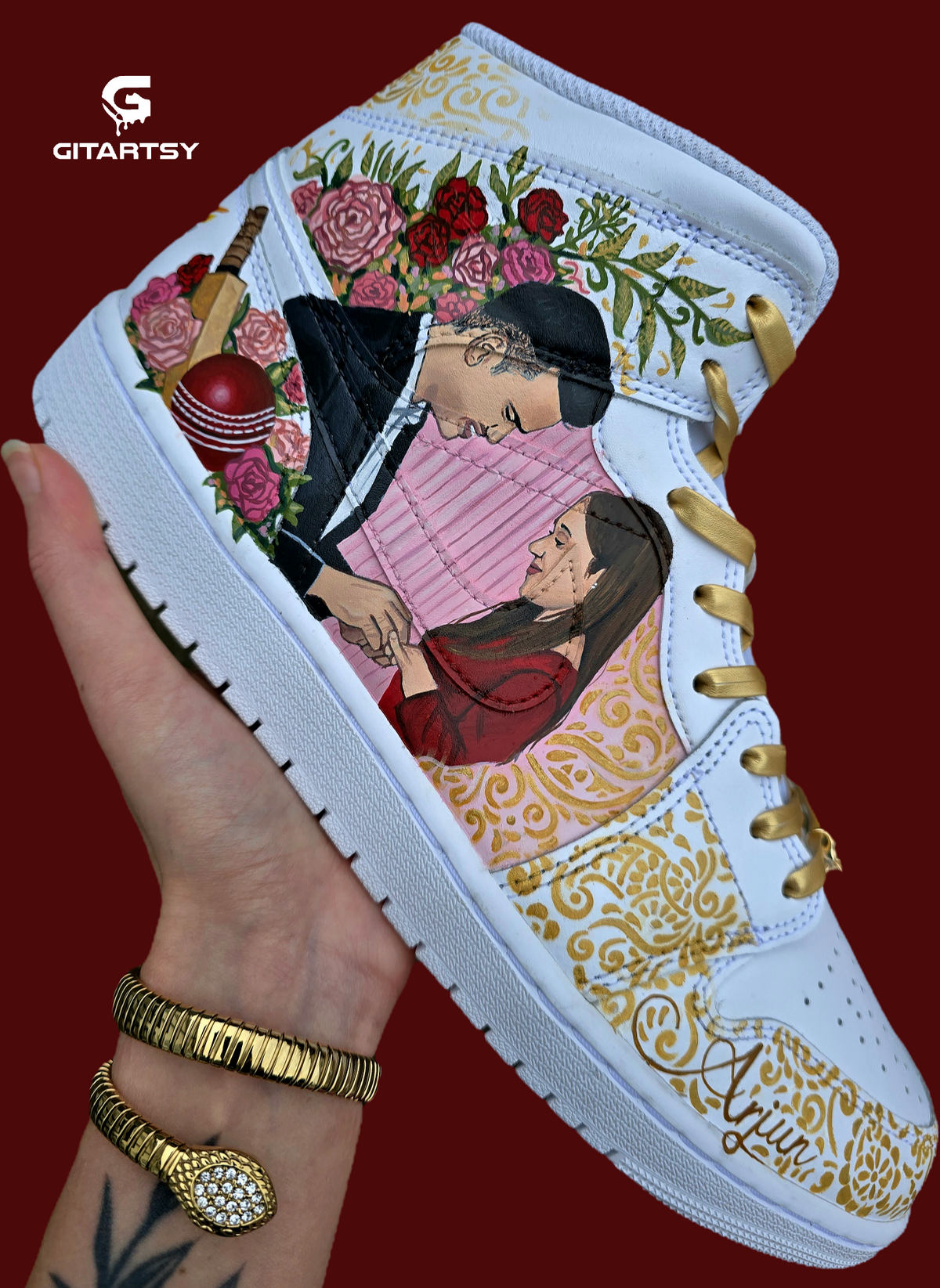 A pair of nike jordan 1 white high-top sneakers with custom illustrations of a couple Arjun Tendulkar and Saaniya Chandhok with gold paisley patterns henna, pets, florals with maroon and blue colour scheme displayed against a dark red background with a GITARTSY logo in the corner.