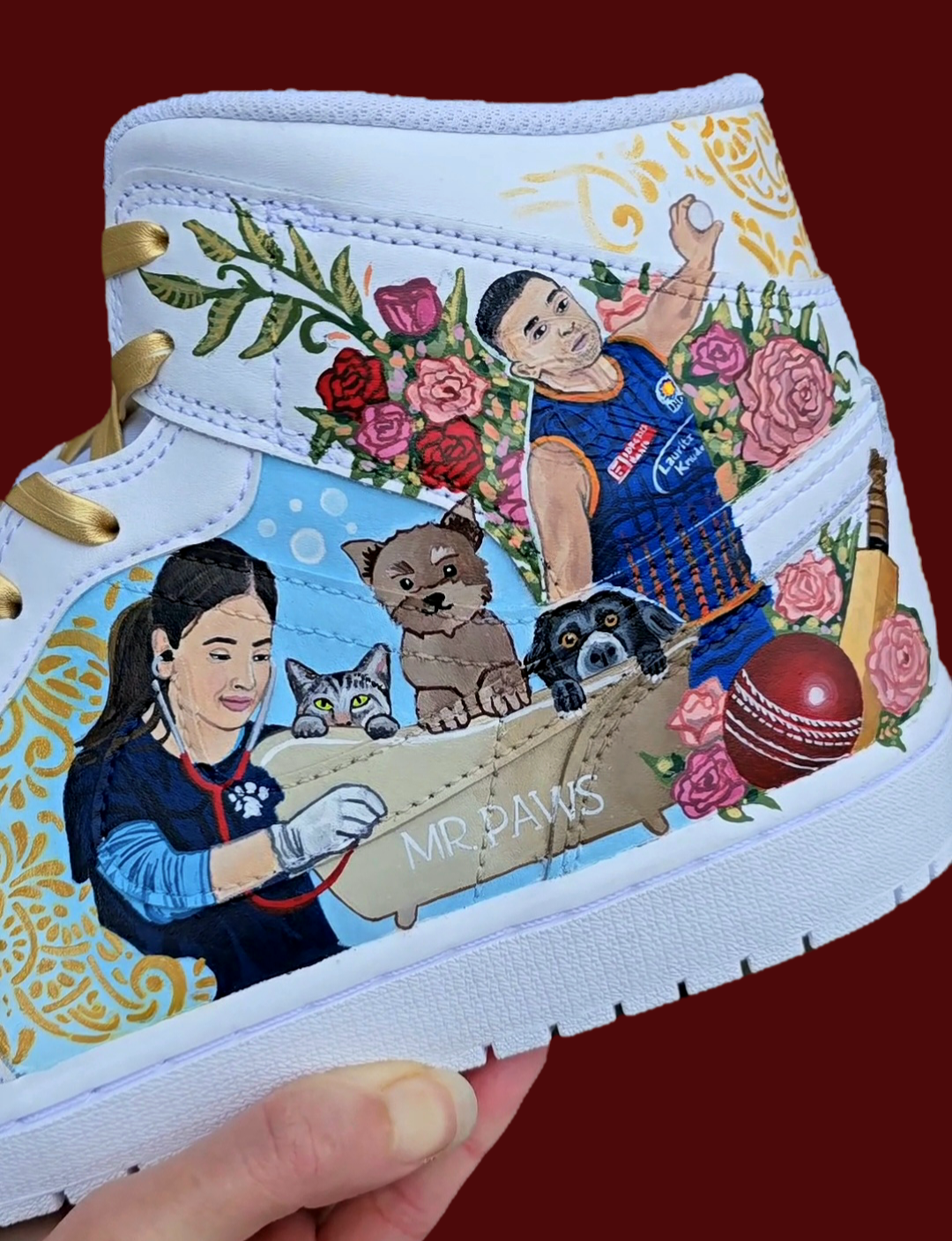 A pair of nike jordan 1 white high-top sneakers with custom illustrations of a couple Arjun Tendulkar and Saaniya Chandhok with gold paisley patterns henna, pets, florals with maroon and blue colour scheme displayed against a dark red background with a GITARTSY logo in the corner.