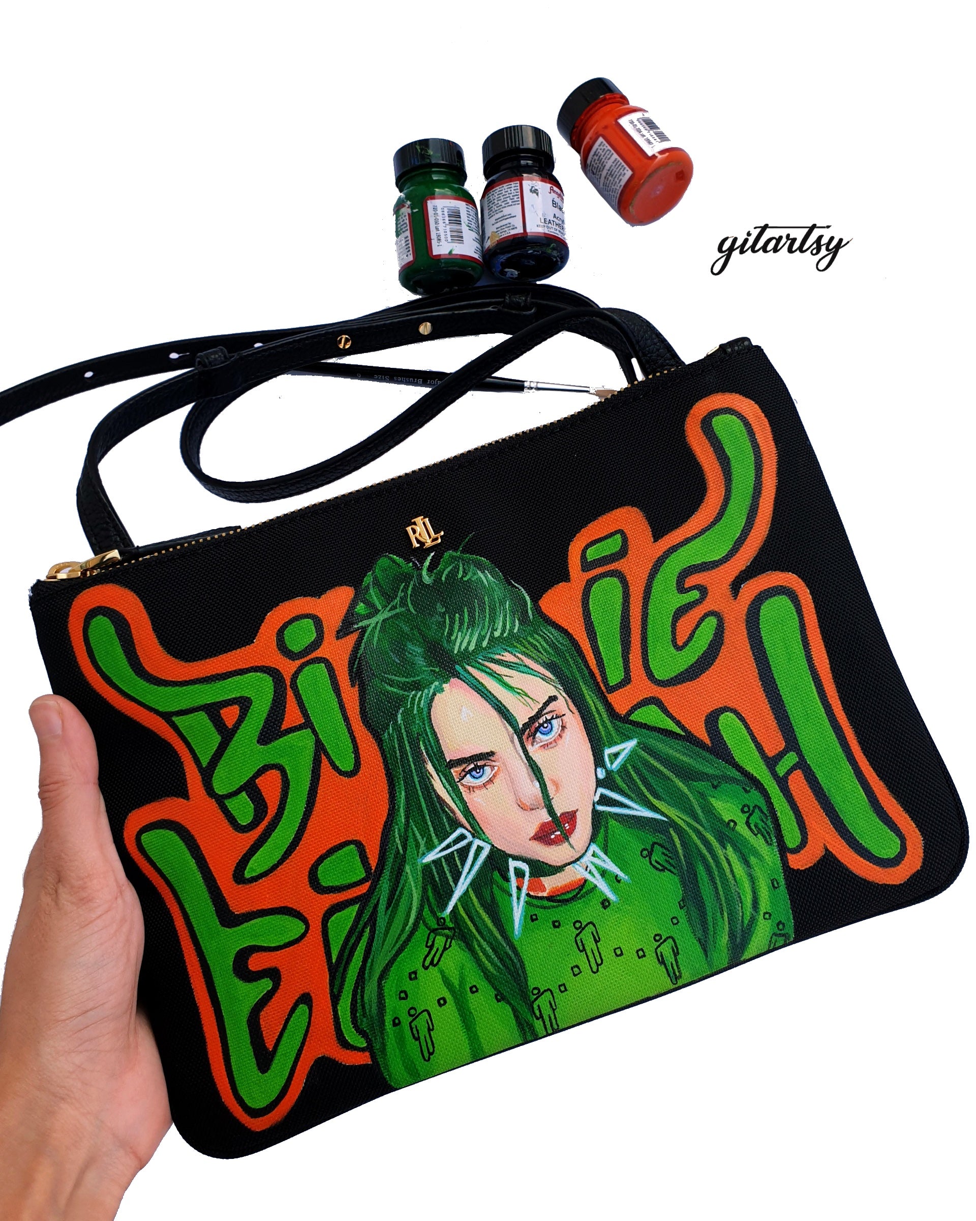 A black Ralph Lauren crossbody canvas bag with a vivid hand-painted portrait of Billie Eilish featuring neon green hair and piercing blue eyes. Surrounding her are graffiti-style green and orange letters spelling “Billie Eilish”. The bag includes a sleek black strap and gold Ralph Lauren logo detailing. High-quality acrylic leather paints are shown in the background alongside a fine detail paintbrush, highlighting the hand-painted craftsmanship.