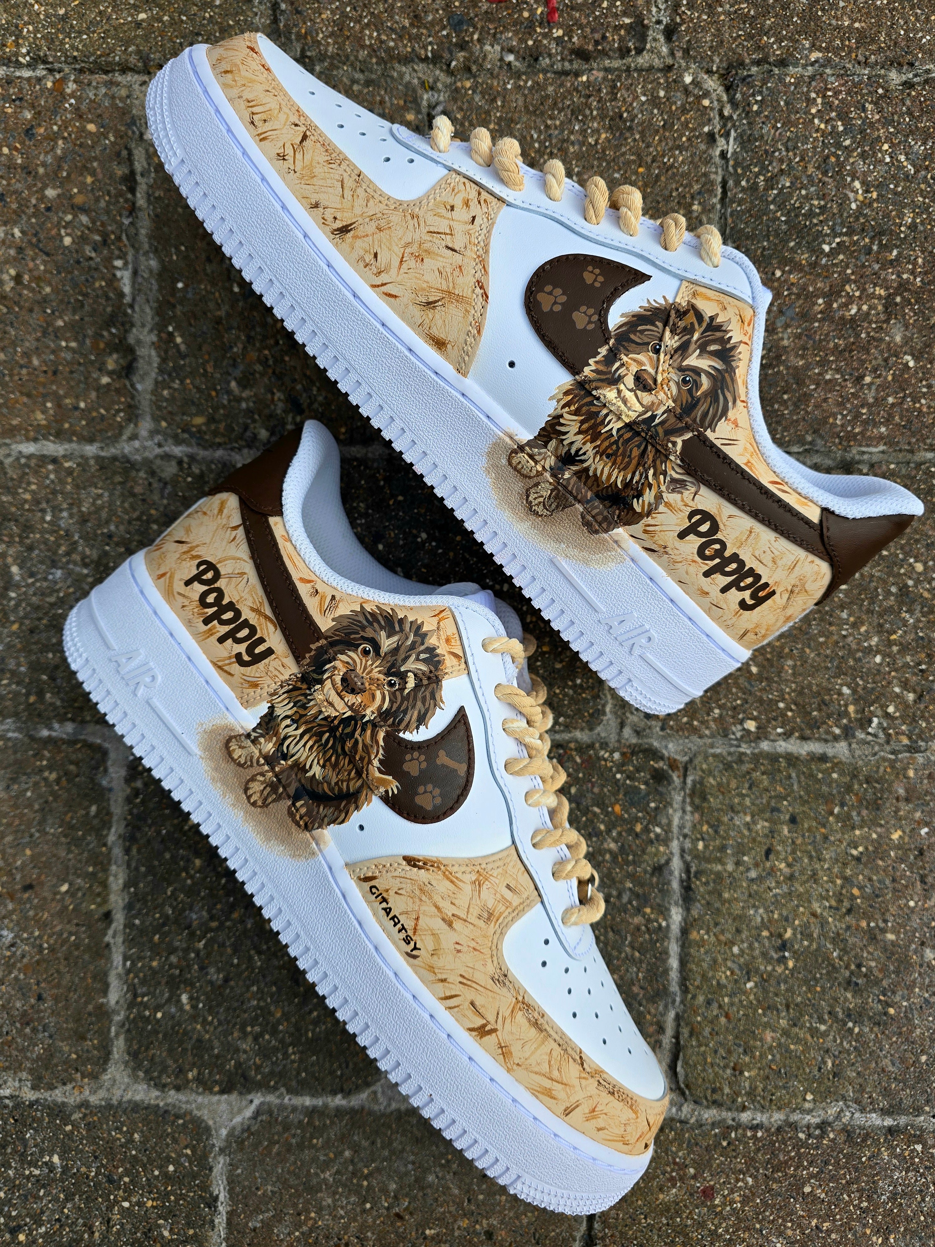Custom hand-painted Nike Air Force 1 sneakers featuring a detailed portrait of a fluffy brown dog named Poppy 🐶. The shoes have a white leather base with light brown textured paint accents and a rich chocolate-brown swoosh adorned with paw prints 🐾. The pet’s name is written in a playful font, and tan rope laces add a rustic touch. Perfect for showcasing love for your pet in a stylish and artistic way.


