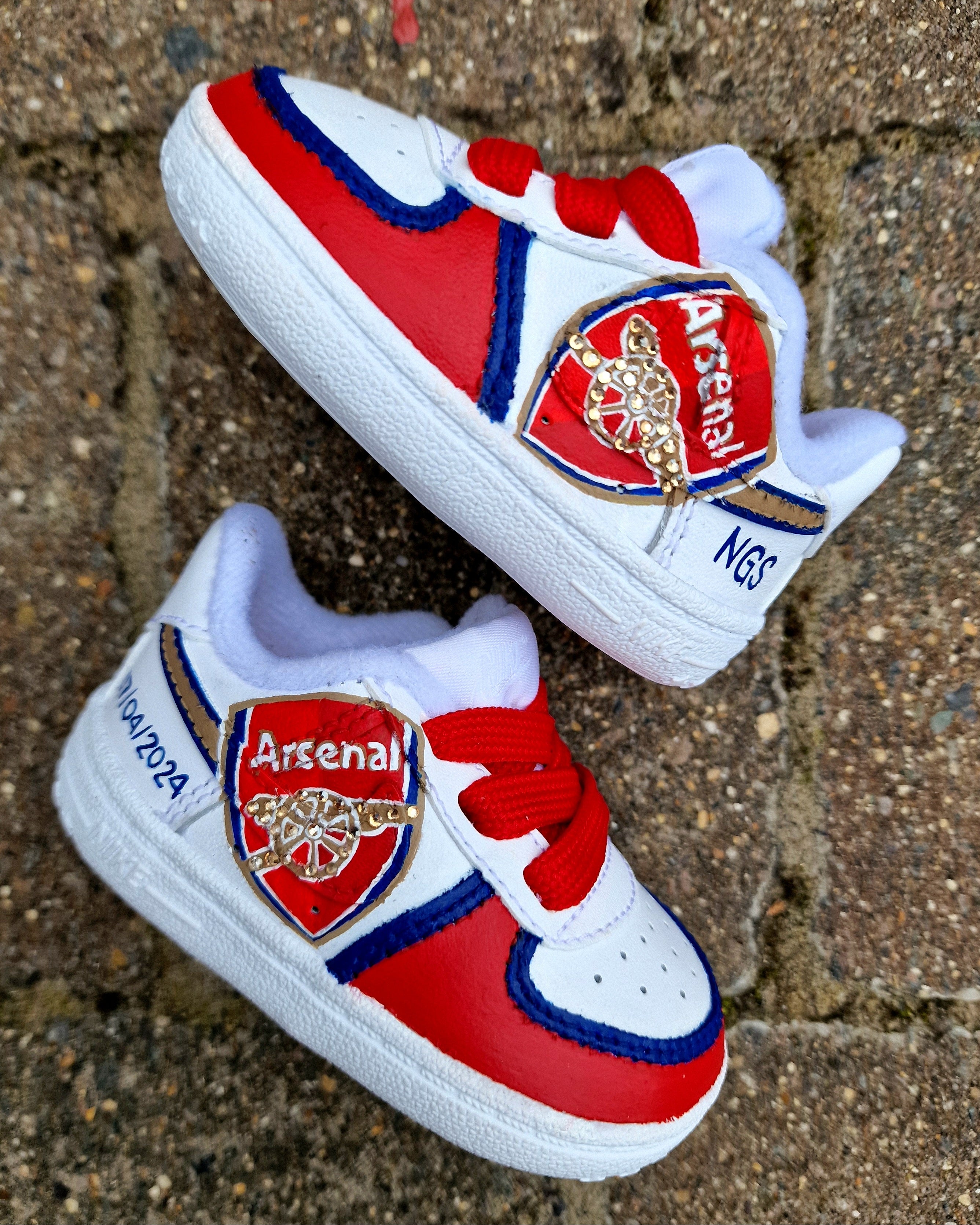 Hand-painted custom Arsenal baby sneakers featuring the official club crest, red and blue colourway, gold accents, personalised name and birth details on white Nike baby shoes. Unique baby gift, football fan keepsake and first shoes for Arsenal supporters. Baby-sized football trainers hand-painted and waterproofed.

