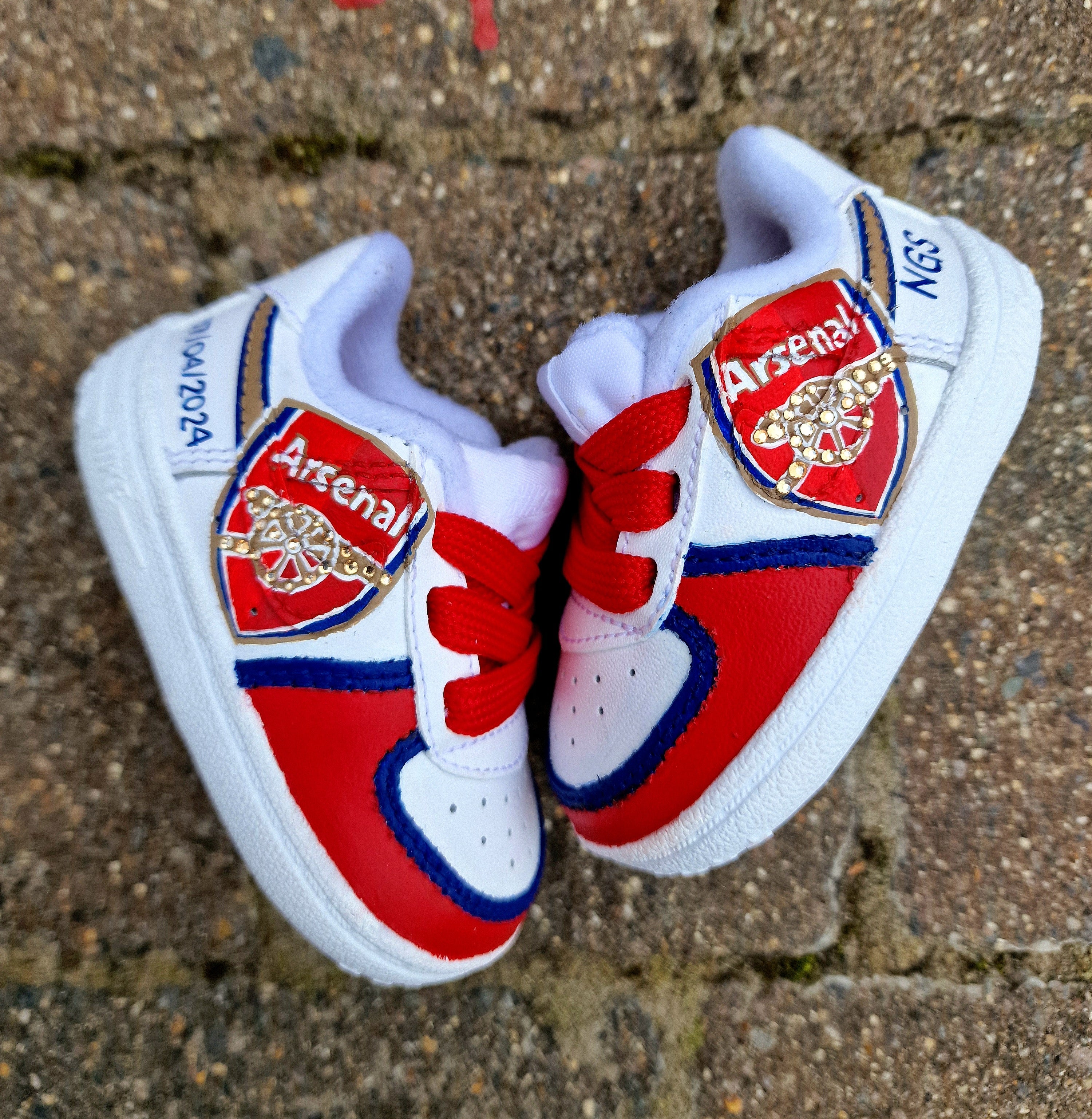 Hand-painted custom Arsenal baby sneakers featuring the official club crest, red and blue colourway, gold accents, personalised name and birth details on white Nike baby shoes. Unique baby gift, football fan keepsake and first shoes for Arsenal supporters. Baby-sized football trainers hand-painted and waterproofed.

