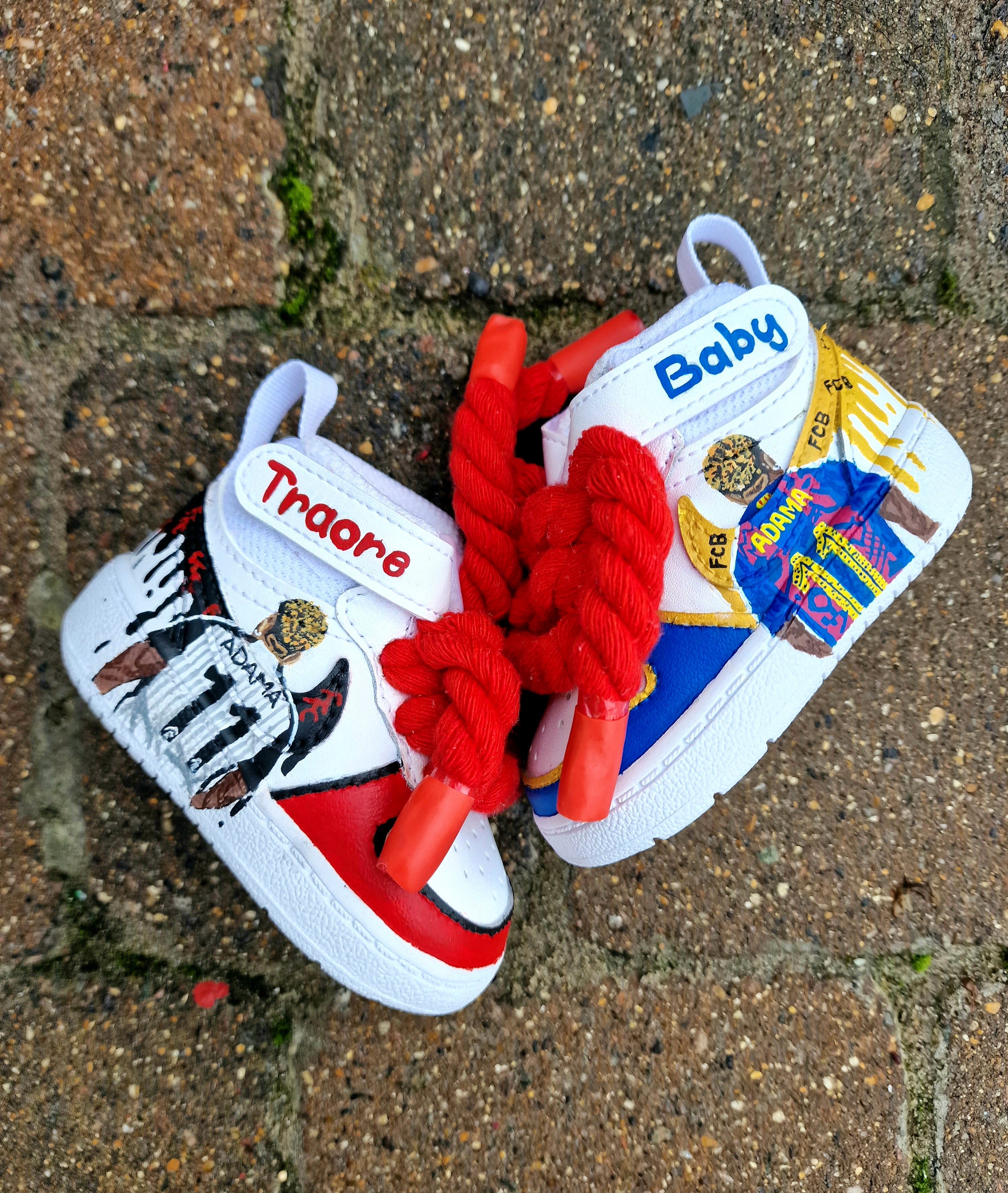 Custom hand-painted baby sneakers inspired by footballer Adama Traoré, featuring FC Barcelona and Fulham FC themes, complete with team logos, jersey illustrations, red braided laces, and personalized strap names on a white baby shoe base.

