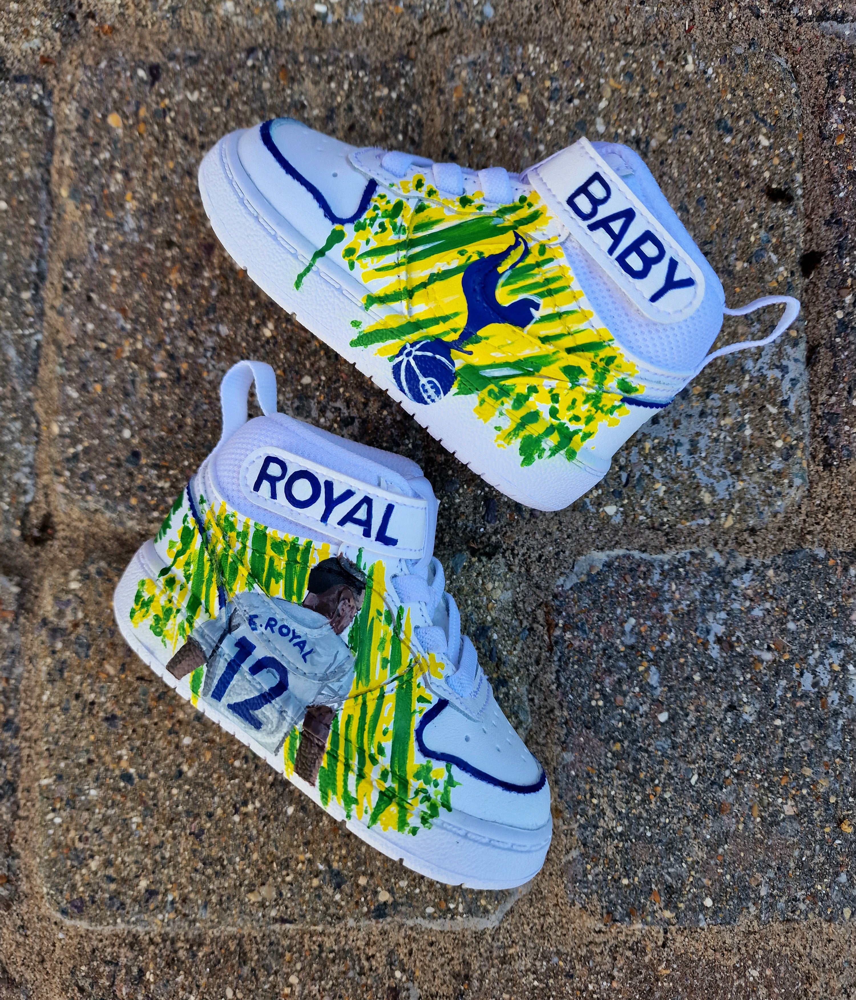 Custom hand-painted baby Adidas sneakers inspired by Brazilian footballer Emerson Royal. Features toddler-sized high-top shoes with illustrations of Emerson in Spurs and Brazil kits, Brazil flag background, colorful 3-stripes, and "Emerson Royal" detailing on the heel. Designed for toddlers, crafted with non-toxic paint, and finished with vibrant detailing by Gitartsy.

