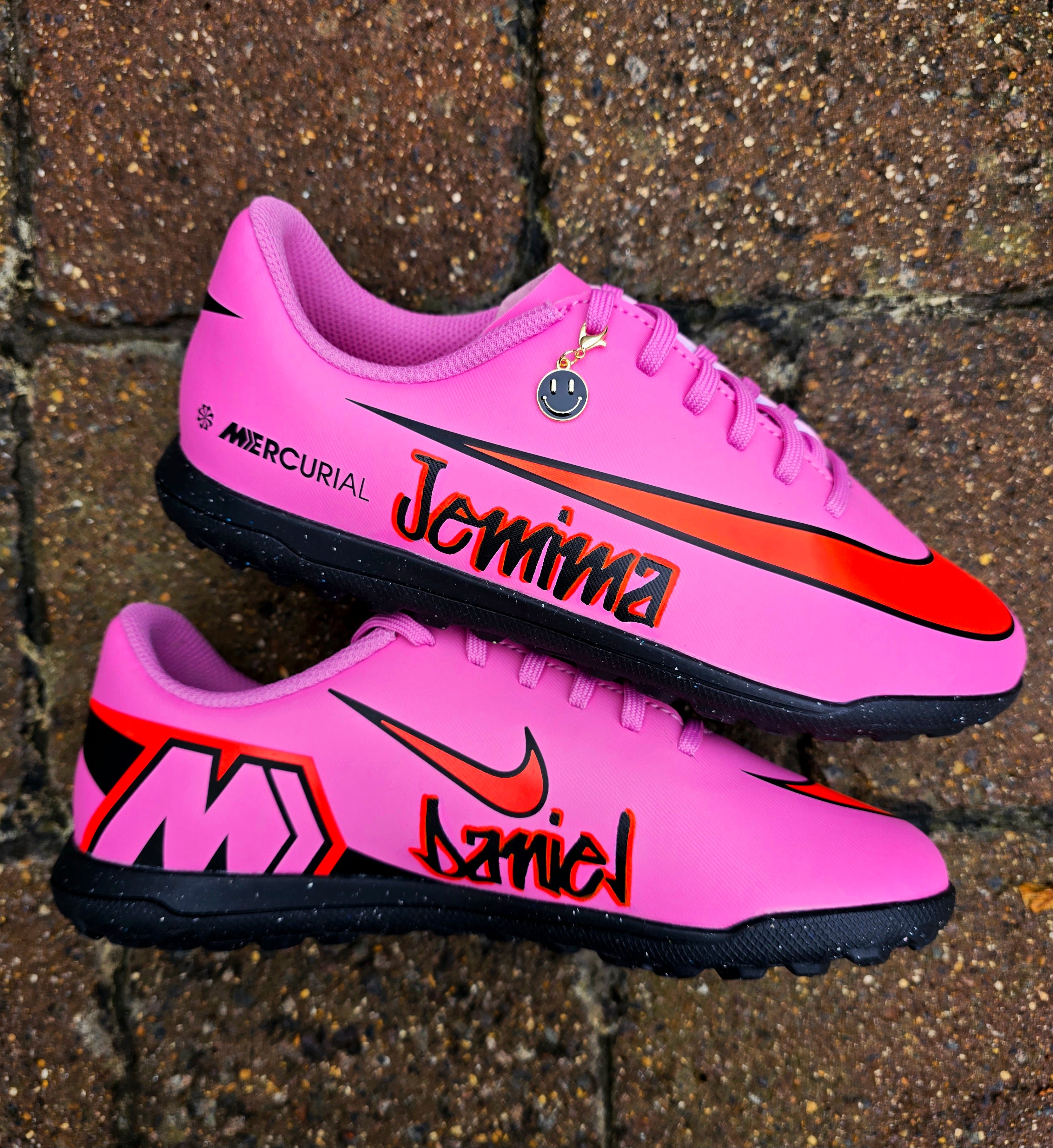 Custom pink Nike Mercurial football sneakers with graffiti-style personalised names in bold red and black lettering. Features include a striking red Nike swoosh, black outsole, and a gold smiley face lace charm. Designed for turf and indoor football, these shoes combine high-performance sportswear with unique, hand-painted street art customisation. Perfect for athletes and collectors seeking one-of-a-kind football boots.


