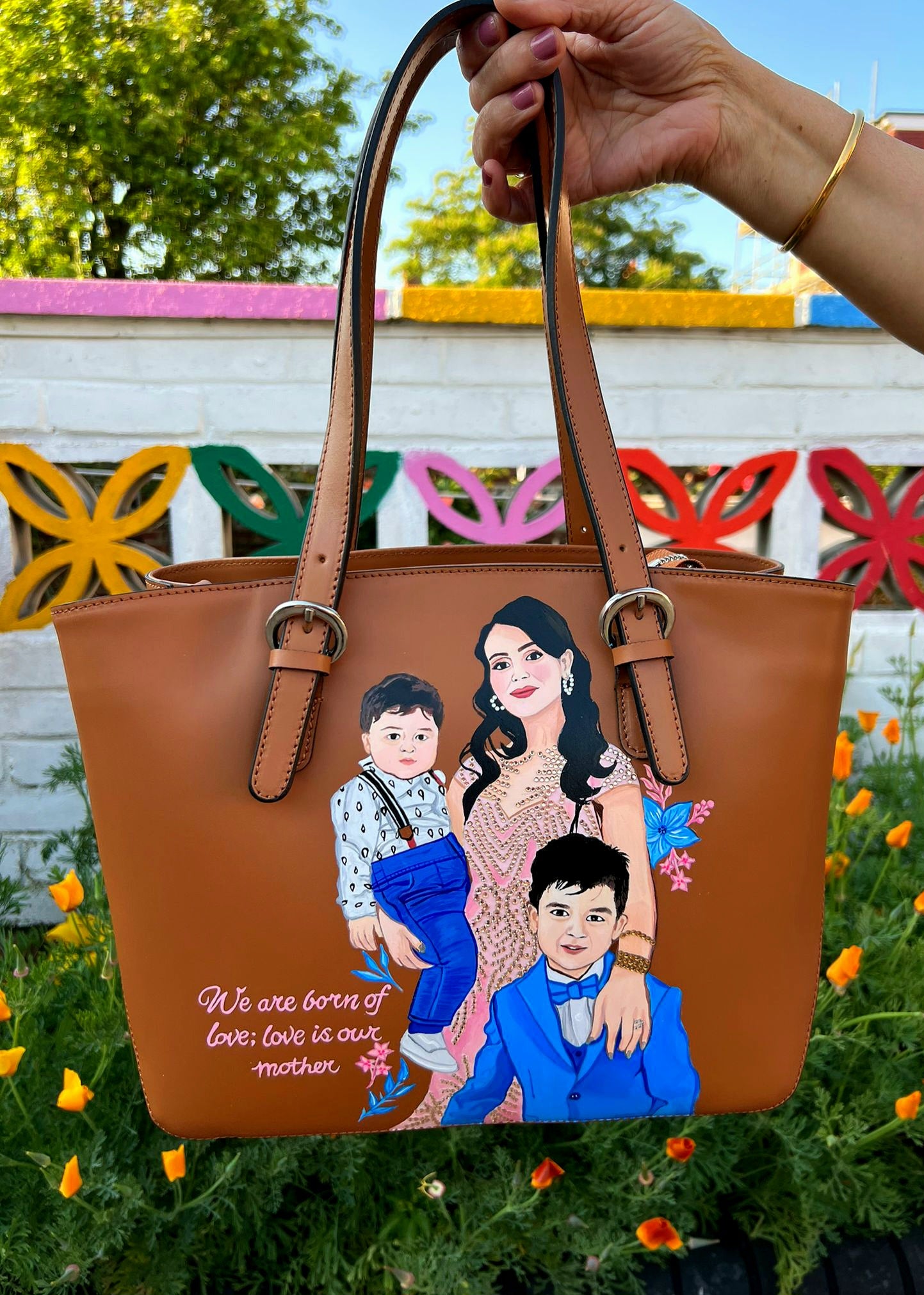 A custom hand-painted tan leather tote bag featuring a family portrait of a mother and two children, designed in a detailed artistic style with floral embellishments and a heartfelt quote. This unique leather bag is personalized from your photos and painted using durable, waterproof paints, ideal as a memory keepsake or meaningful gift for mothers. The painted design stands out against the rich tan leather, making it both functional and sentimental.

