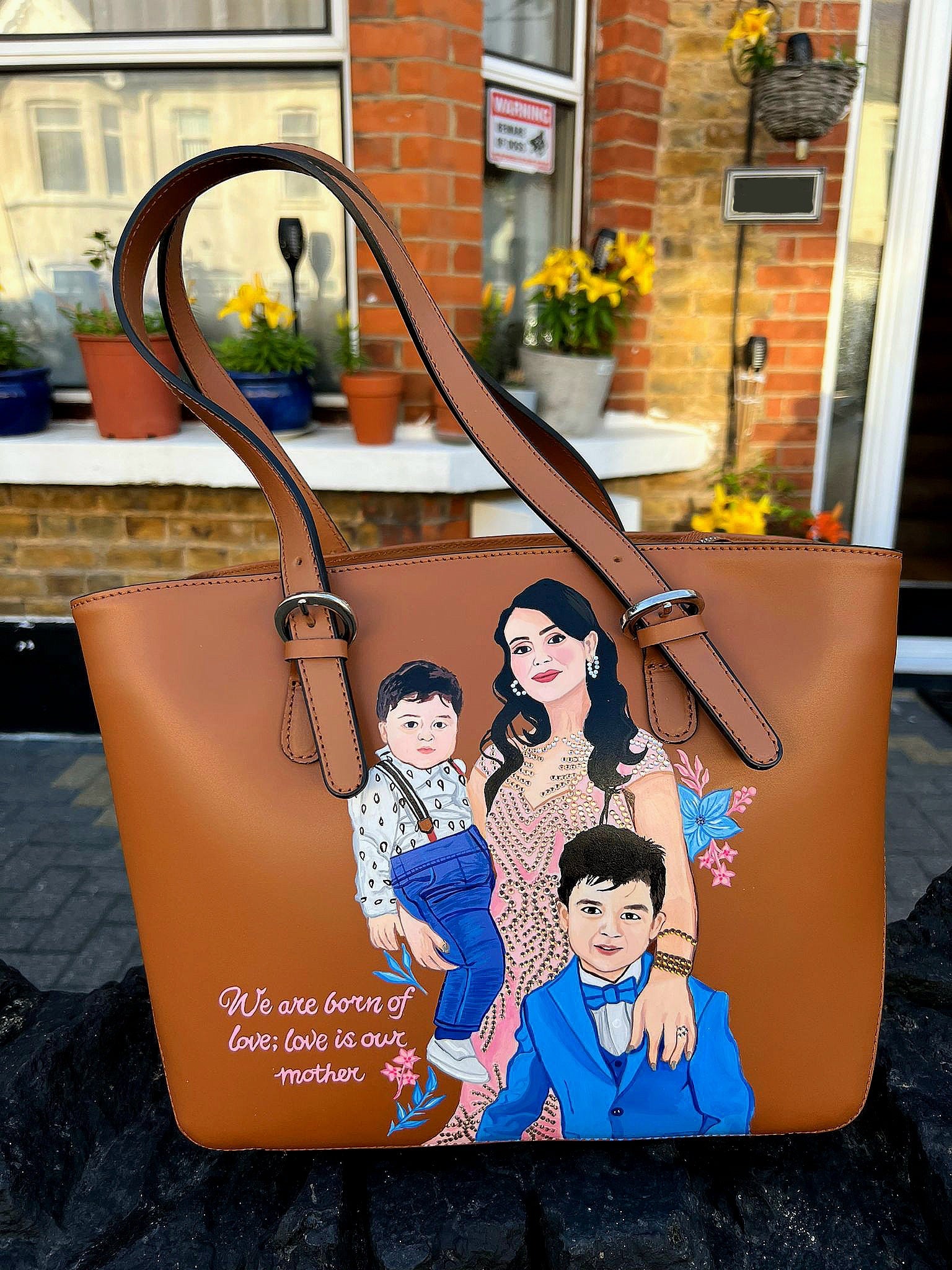 A custom hand-painted tan leather tote bag featuring a family portrait of a mother and two children, designed in a detailed artistic style with floral embellishments and a heartfelt quote. This unique leather bag is personalized from your photos and painted using durable, waterproof paints, ideal as a memory keepsake or meaningful gift for mothers. The painted design stands out against the rich tan leather, making it both functional and sentimental.

