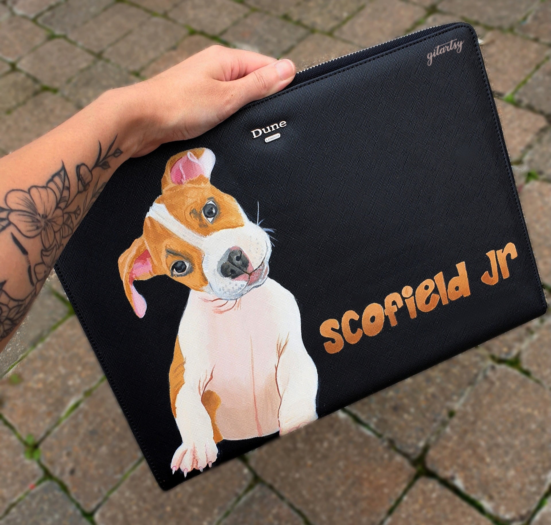 A sleek black Dune London leather pouch featuring a highly detailed hand-painted portrait of a brown and white puppy with pink ears and expressive eyes. The pet’s name “Scofield Jr” is painted in a warm orange graffiti-style font. Angelus leather paints in various shades including salmon, dark taupe, hot pink, and brick are placed beside the pouch, showcasing the artistic process. The pouch has a silver zipper and textured leather finish.

