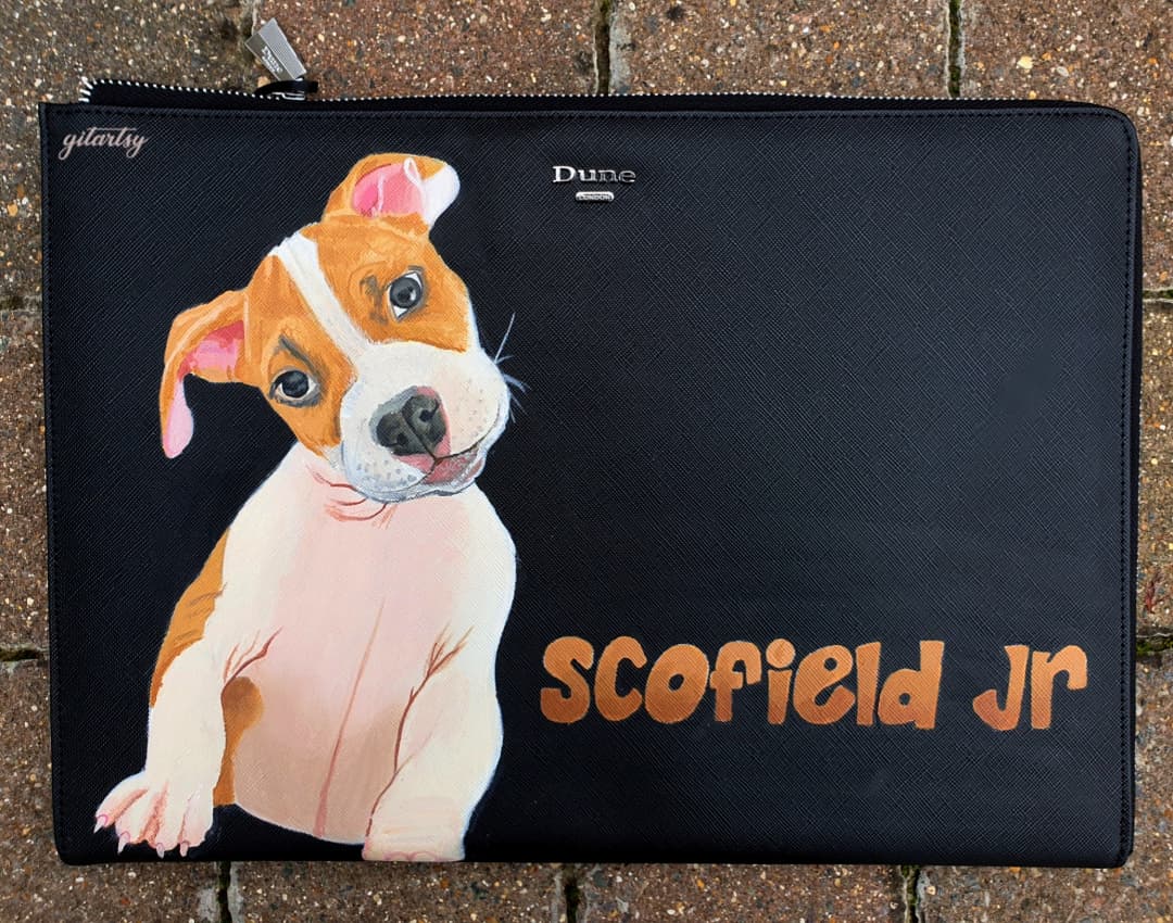 A sleek black Dune London leather pouch featuring a highly detailed hand-painted portrait of a brown and white puppy with pink ears and expressive eyes. The pet’s name “Scofield Jr” is painted in a warm orange graffiti-style font. Angelus leather paints in various shades including salmon, dark taupe, hot pink, and brick are placed beside the pouch, showcasing the artistic process. The pouch has a silver zipper and textured leather finish.

