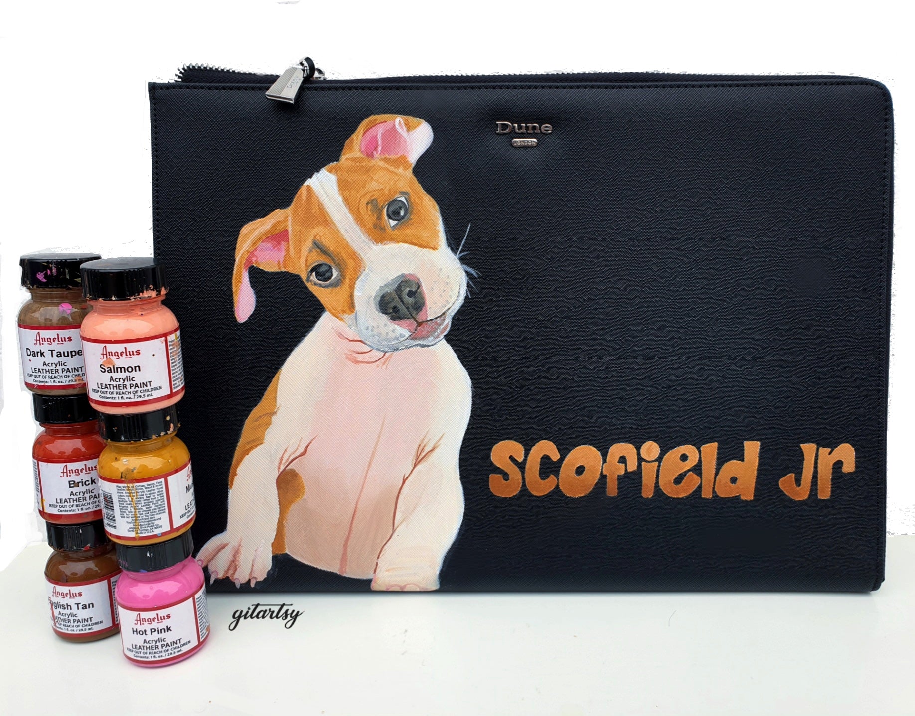 A sleek black Dune London leather pouch featuring a highly detailed hand-painted portrait of a brown and white puppy with pink ears and expressive eyes. The pet’s name “Scofield Jr” is painted in a warm orange graffiti-style font. Angelus leather paints in various shades including salmon, dark taupe, hot pink, and brick are placed beside the pouch, showcasing the artistic process. The pouch has a silver zipper and textured leather finish.

