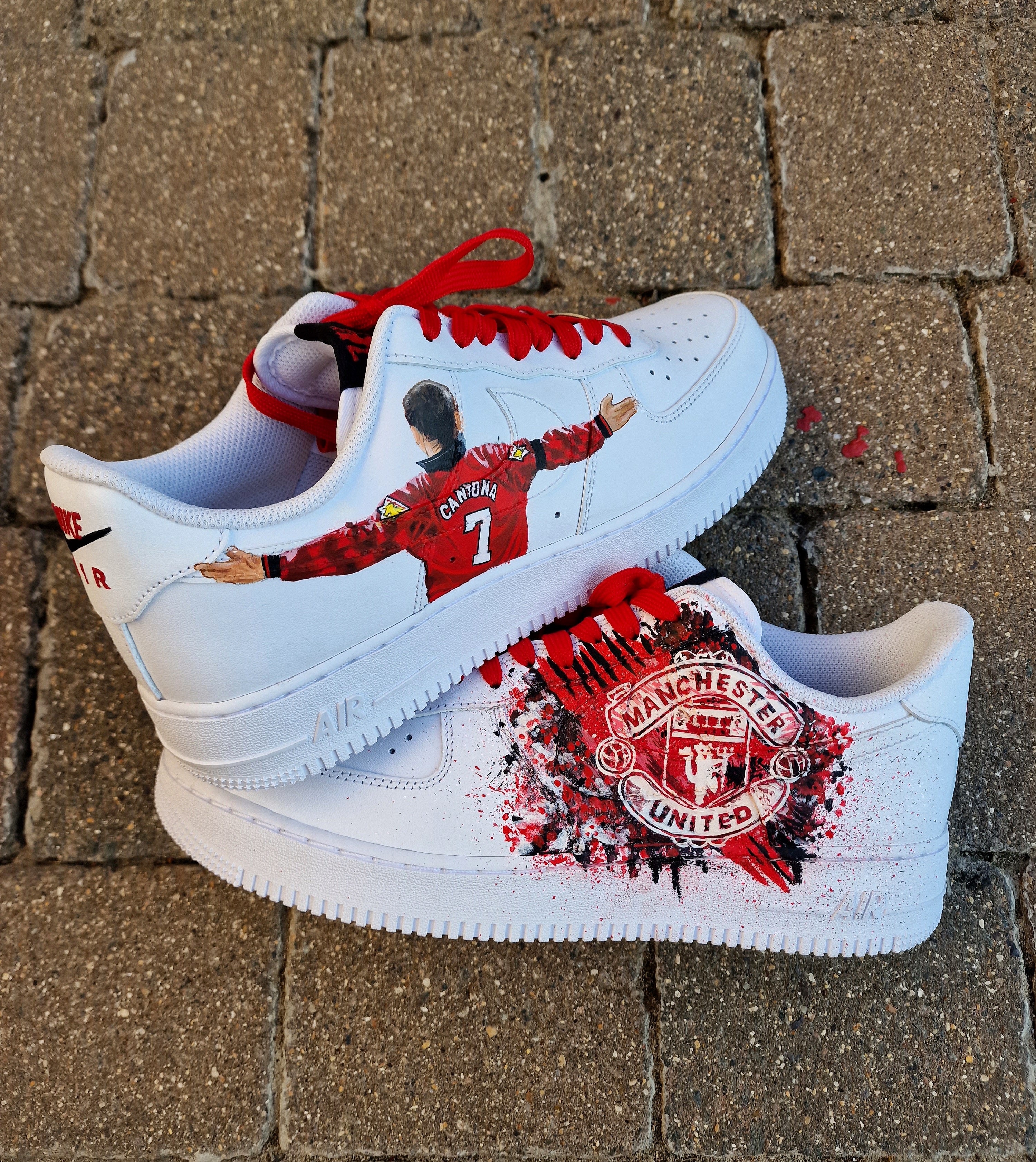 Custom Nike Air Force 1 sneakers hand-painted with Manchester United's club crest on one side in bold red-black splatter, and Eric Cantona's famous goal celebration pose with arms outstretched wearing the No. 7 jersey on the opposite side. Finished with red laces and premium leather base, these AF1s are waterproof-coated and crafted with attention to detail, offering a perfect gift for Manchester United fans.

