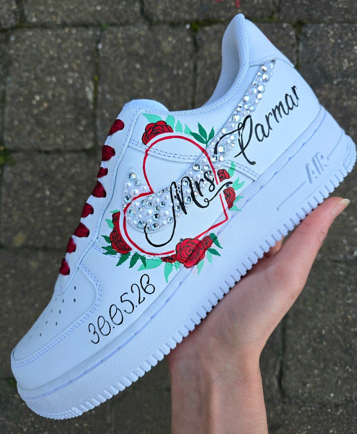 A hand holds a white custom sneaker featuring a red heart outline, rhinestones, red roses, and the text 'Mrs. Parmar' and '30. bridal custom sneaker