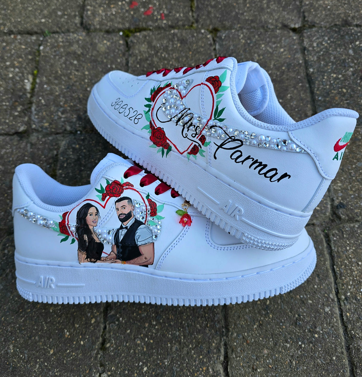A pair of white custom Nike Air Force 1 sneakers displayed on a gray stone pavement. The shoes are decorated with red roses, rhinestones, custom photo prints of a couple, and the text 'Mrs.