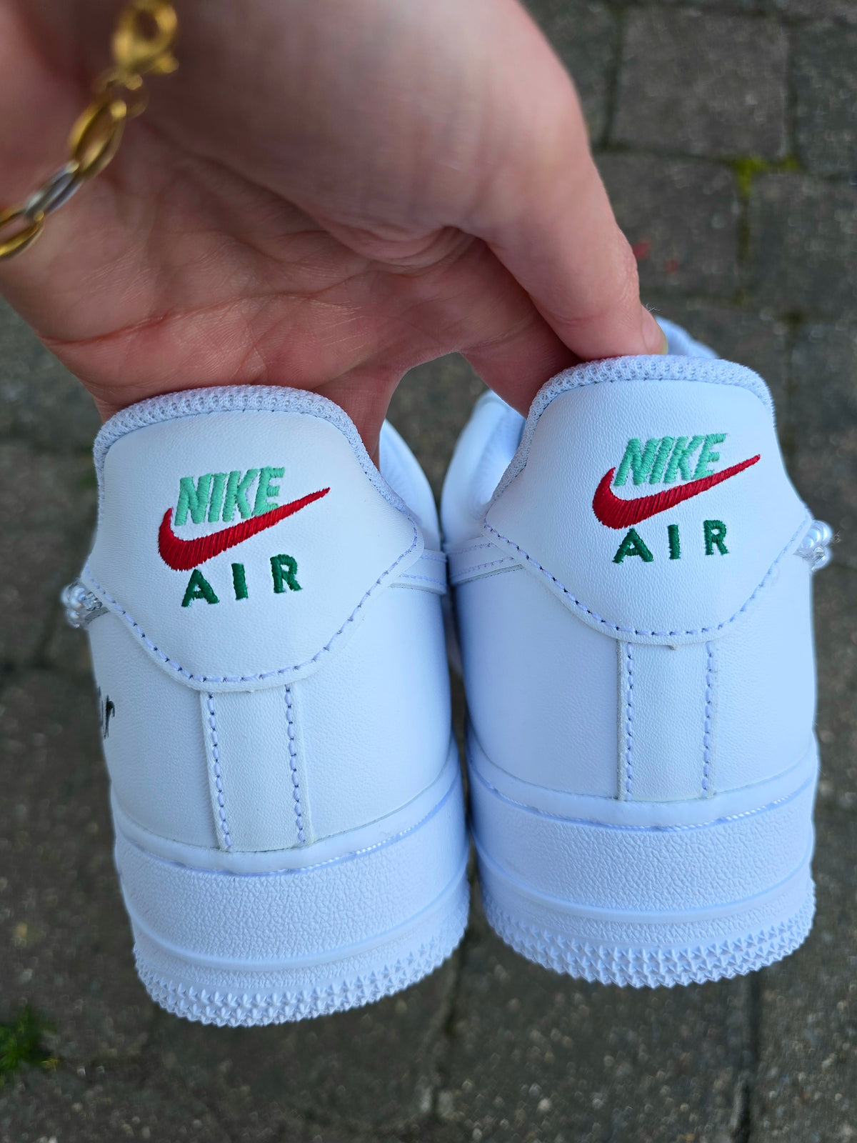A pair of white Nike Air Force 1 Low sneakers shown from the back, held by a hand over a gray paved ground. The tongue of each shoe features the green 'NIKE AIR' text and red Swoosh logo.