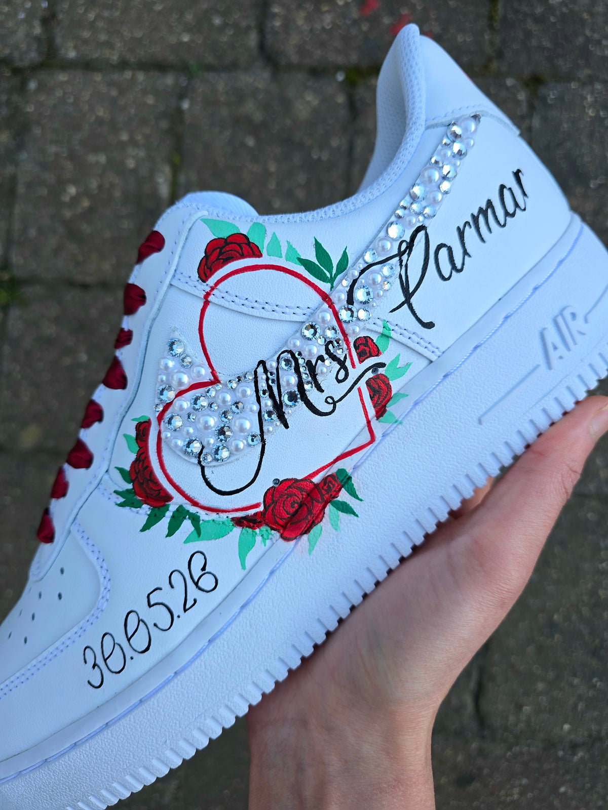 A white custom-painted Nike Air Force 1 sneaker held in a hand, decorated with red roses, green leaves, a red heart outline, rhinestones, and black script text reading 'Mrs. Parmar' and '306528'.