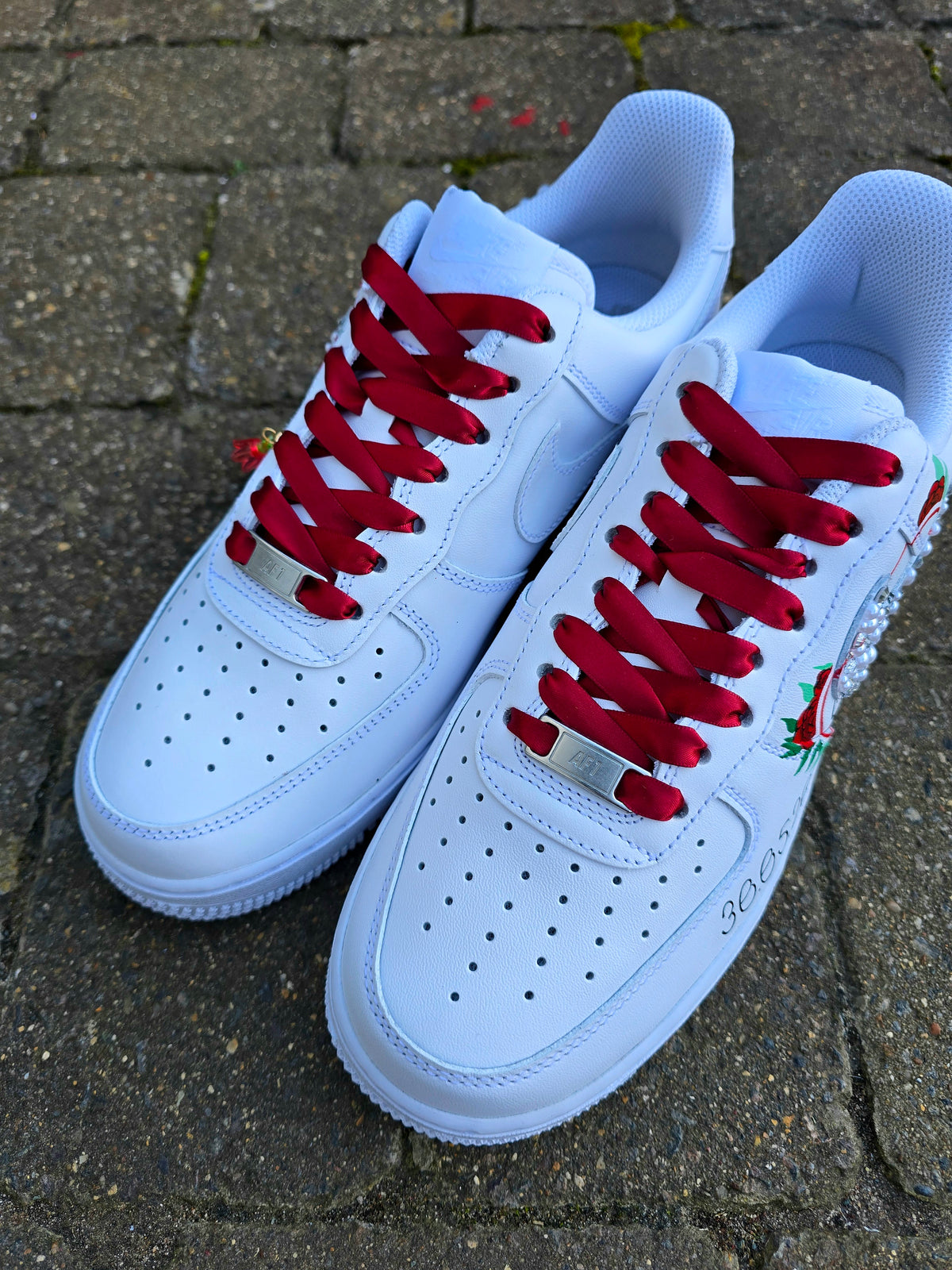A pair of white low-top sneakers with bright red laces, placed on a gray stone pavement. The shoes feature perforated detailing on the toe box, a white rubber sole, and a small red and green floral embellishment on the side.