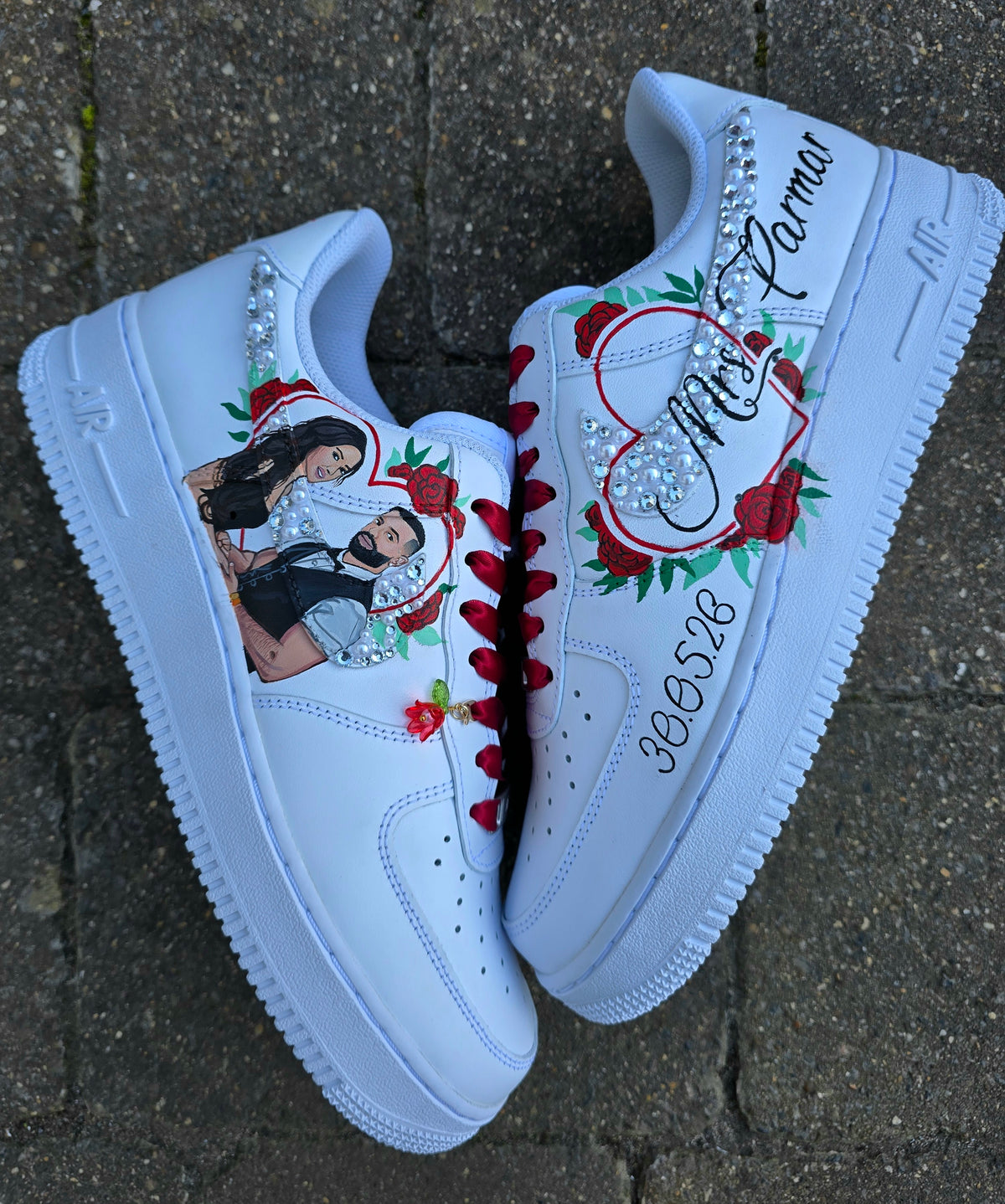 A pair of white custom-painted Nike Air Force 1-style sneakers placed on a gray stone surface. The shoes feature red embroidered laces, red roses, green leaves, rhinestone embellishments, a custom photo print on the left shoe, and the name 'Mrs Parmar' with a heart and number on the right shoe.