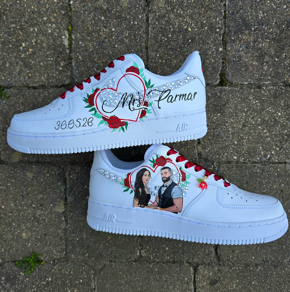 A pair of white custom Air Force 1-style sneakers placed on a gray stone pavement. The shoes feature hand-painted red roses, a glittery heart, a couple's portrait, rhinestone embellishments, and custom text including 'Mrs.