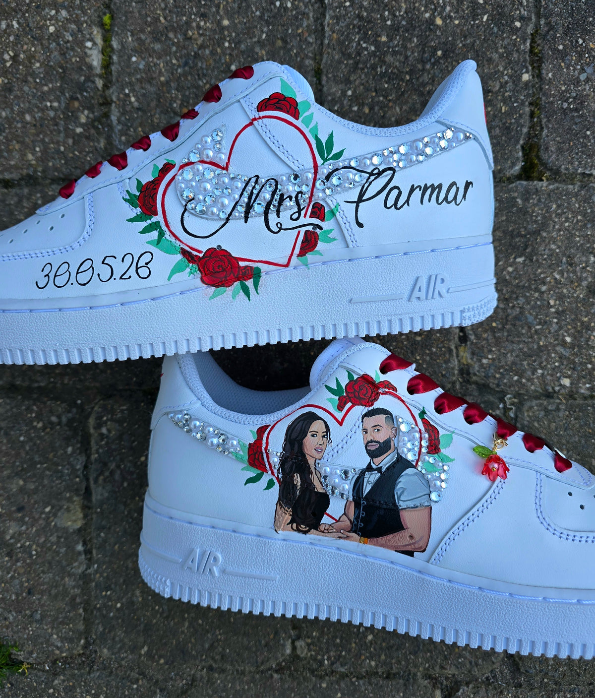 A pair of white custom Air Force 1-style sneakers with red laces, a large red heart outline, rhinestones, roses, and handwritten text on the sides, displayed on a gray stone surface.