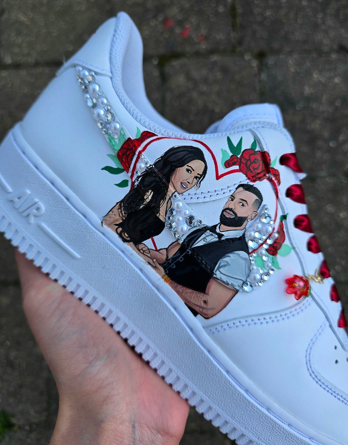 A white sneaker held in a hand, decorated with a custom illustration of a couple inside a heart, red roses, and rhinestone embellishments, set against a blurred outdoor background.