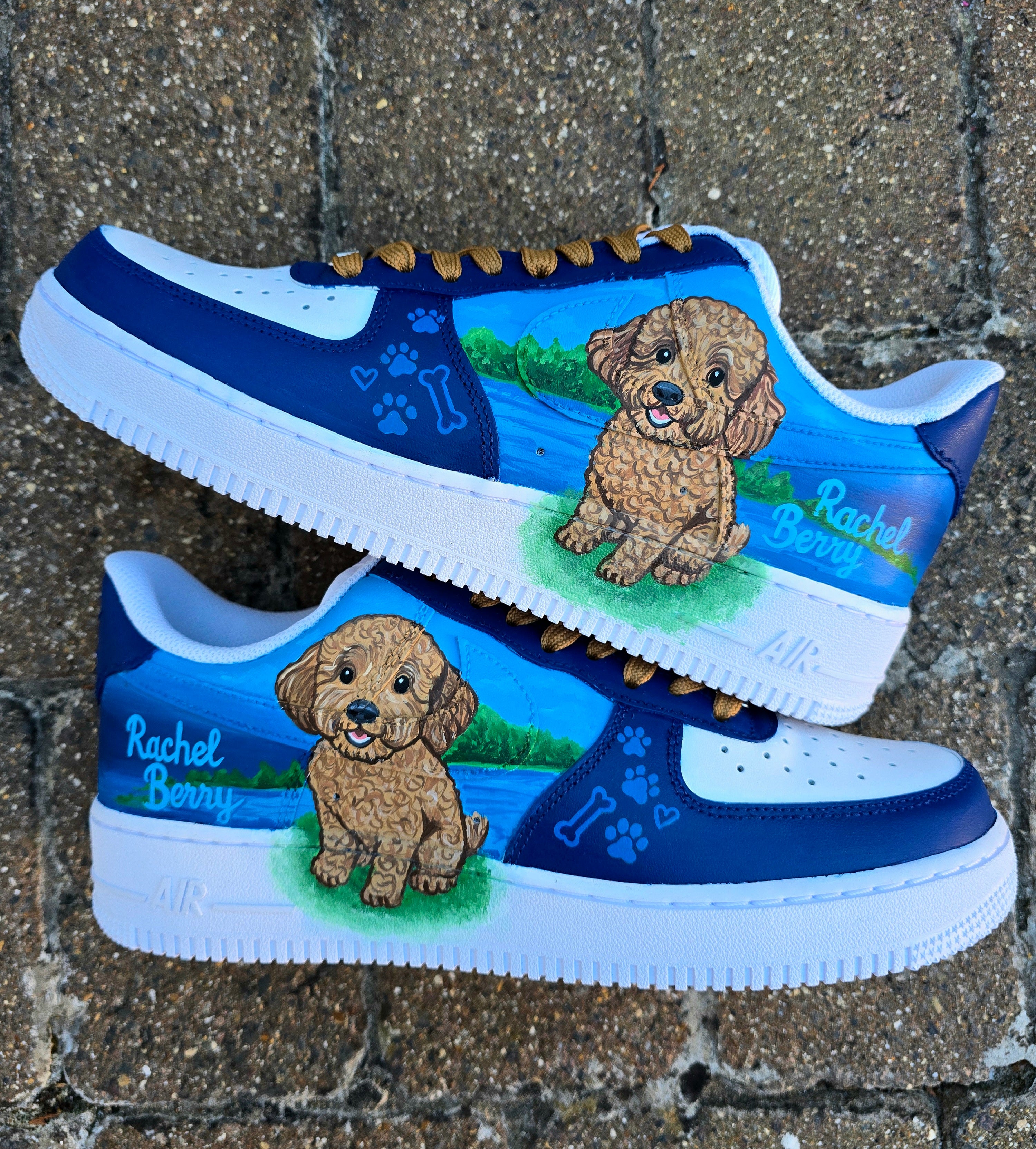 Custom Nike Air Force 1 sneakers 🐾 hand-painted with a detailed toy poodle pet portrait, featuring a caramel-colored curly-haired dog sitting in a grassy field with a blue sky and lake background. Includes the pet’s name in stylish script, paw prints, bones, and heart icons, all painted on a blue and navy color-block base with golden-brown laces. Each pair is personalized to match your pet’s likeness and crafted using durable, waterproof leather paints, making them wearable art for dog lovers.

