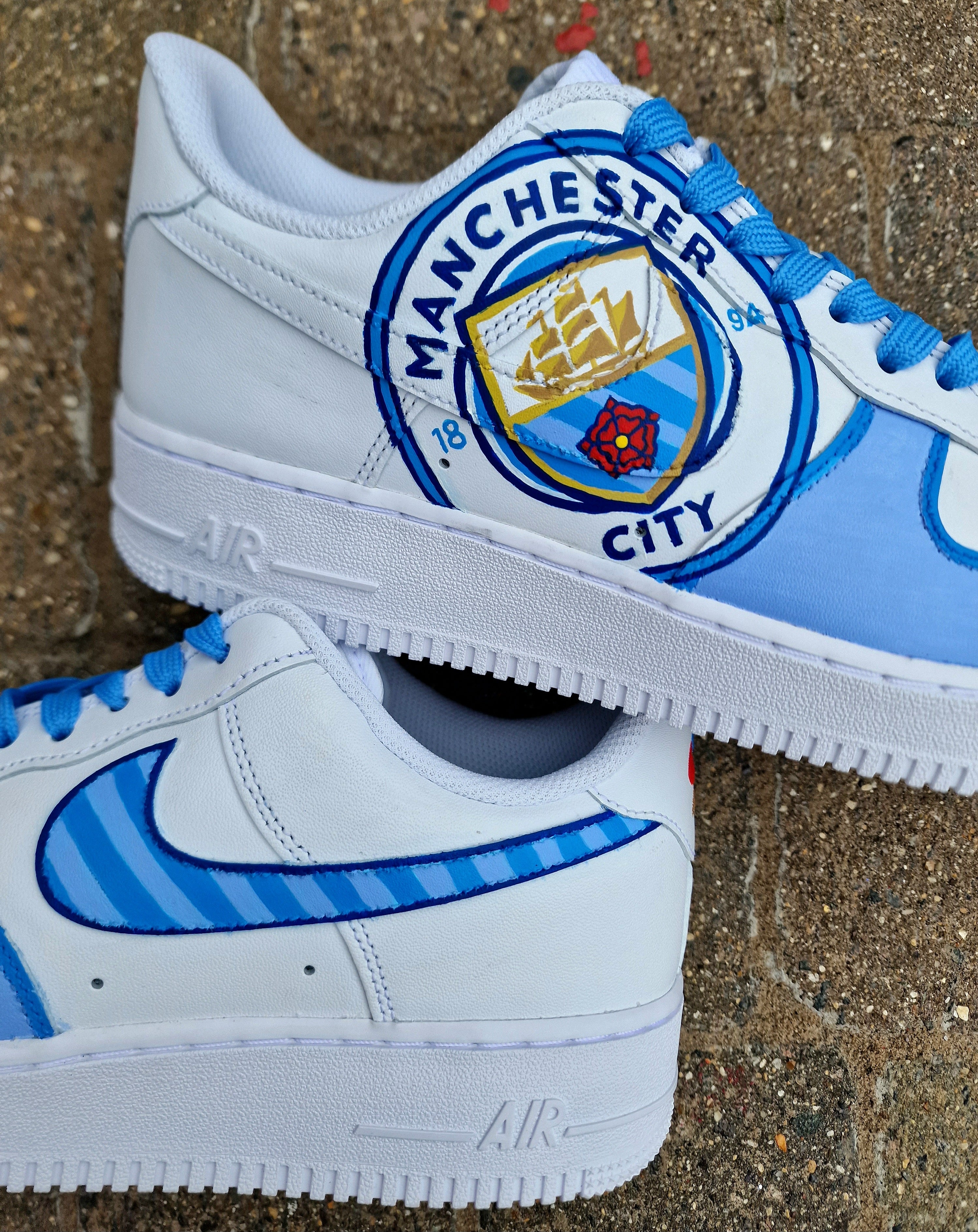 A pair of custom hand-painted Nike Air Force 1 sneakers featuring detailed artwork of the Manchester City Football Club crest, signature blue hues, striped swoosh, and personalized finishing, crafted by Gitartsy for football fans and sneaker collectors.

