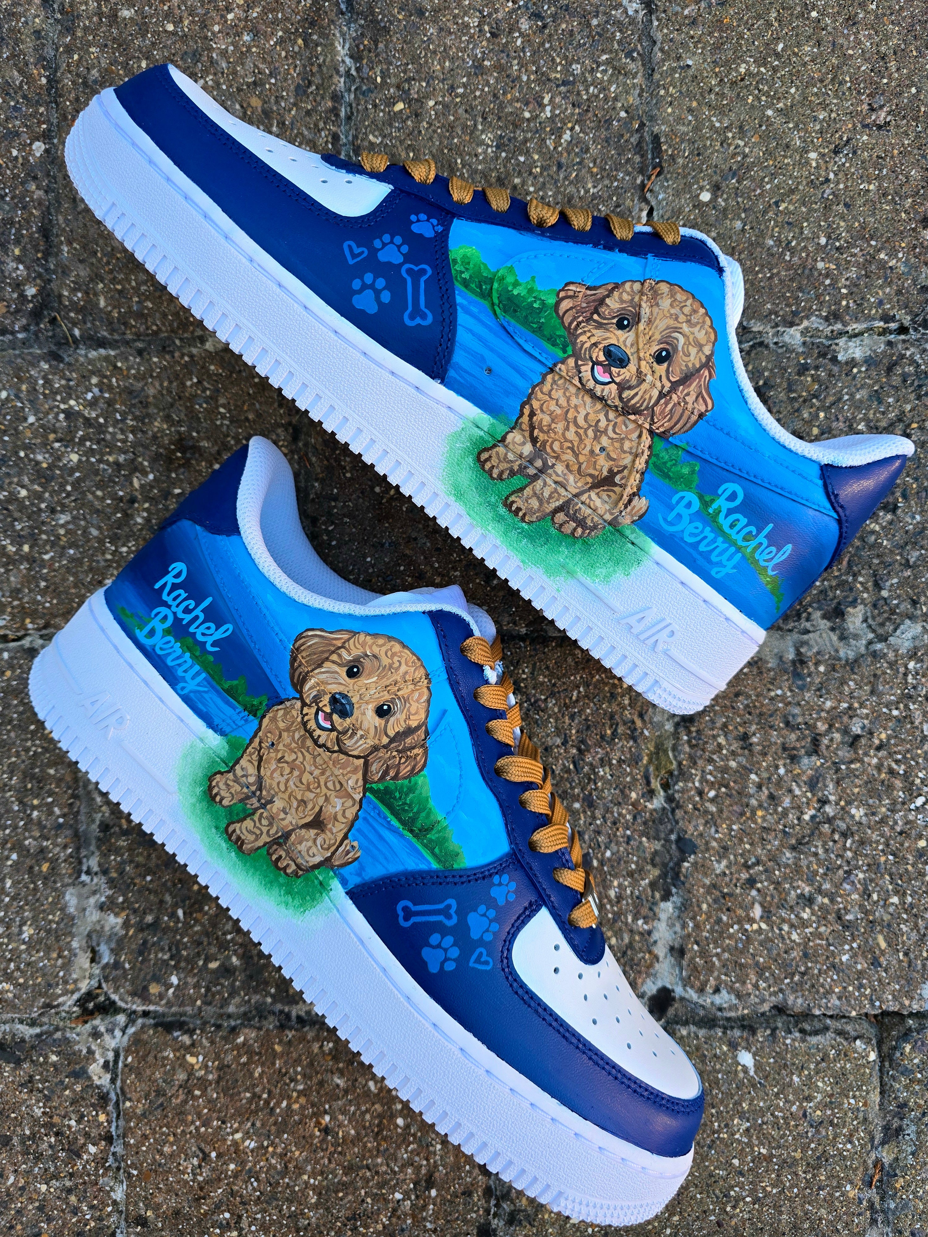 Custom Nike Air Force 1 sneakers 🐾 hand-painted with a detailed toy poodle pet portrait, featuring a caramel-colored curly-haired dog sitting in a grassy field with a blue sky and lake background. Includes the pet’s name in stylish script, paw prints, bones, and heart icons, all painted on a blue and navy color-block base with golden-brown laces. Each pair is personalized to match your pet’s likeness and crafted using durable, waterproof leather paints, making them wearable art for dog lovers.


