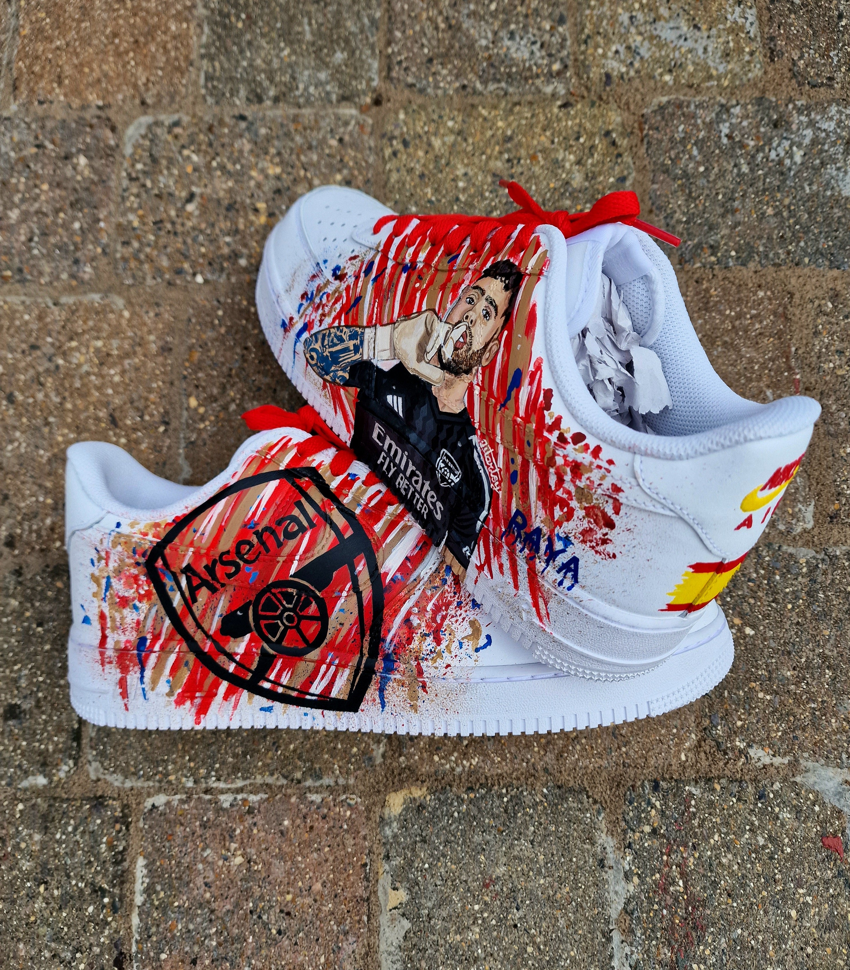 Hand-painted custom Nike Air Force 1 sneakers featuring David Raya in Arsenal goalkeeper kit, Arsenal crest, Spanish flag and colors, vibrant football-inspired splatter artwork, crafted by Gitartsy.


