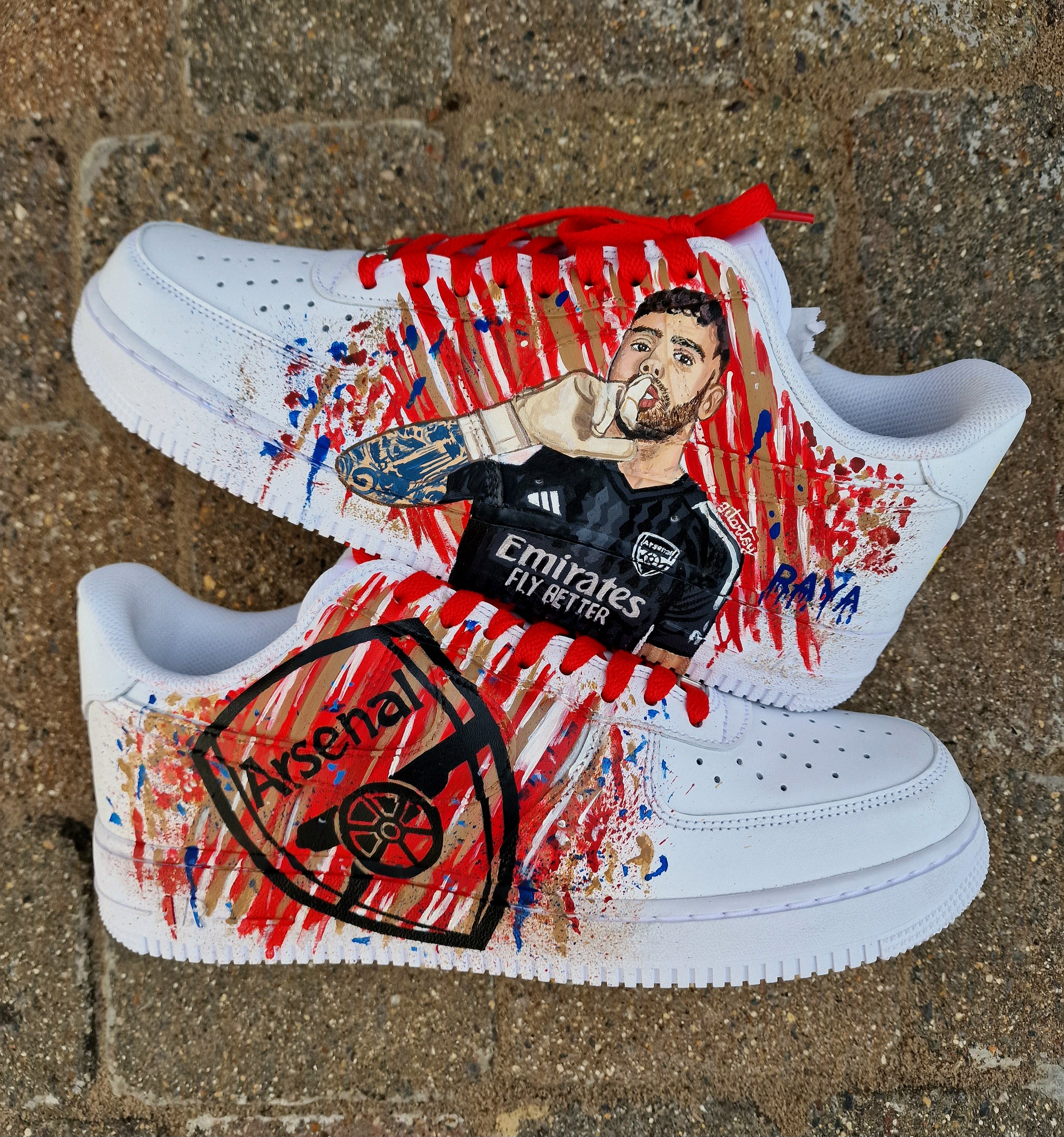Hand-painted custom Nike Air Force 1 sneakers featuring David Raya in Arsenal goalkeeper kit, Arsenal crest, Spanish flag and colors, vibrant football-inspired splatter artwork, crafted by Gitartsy.

