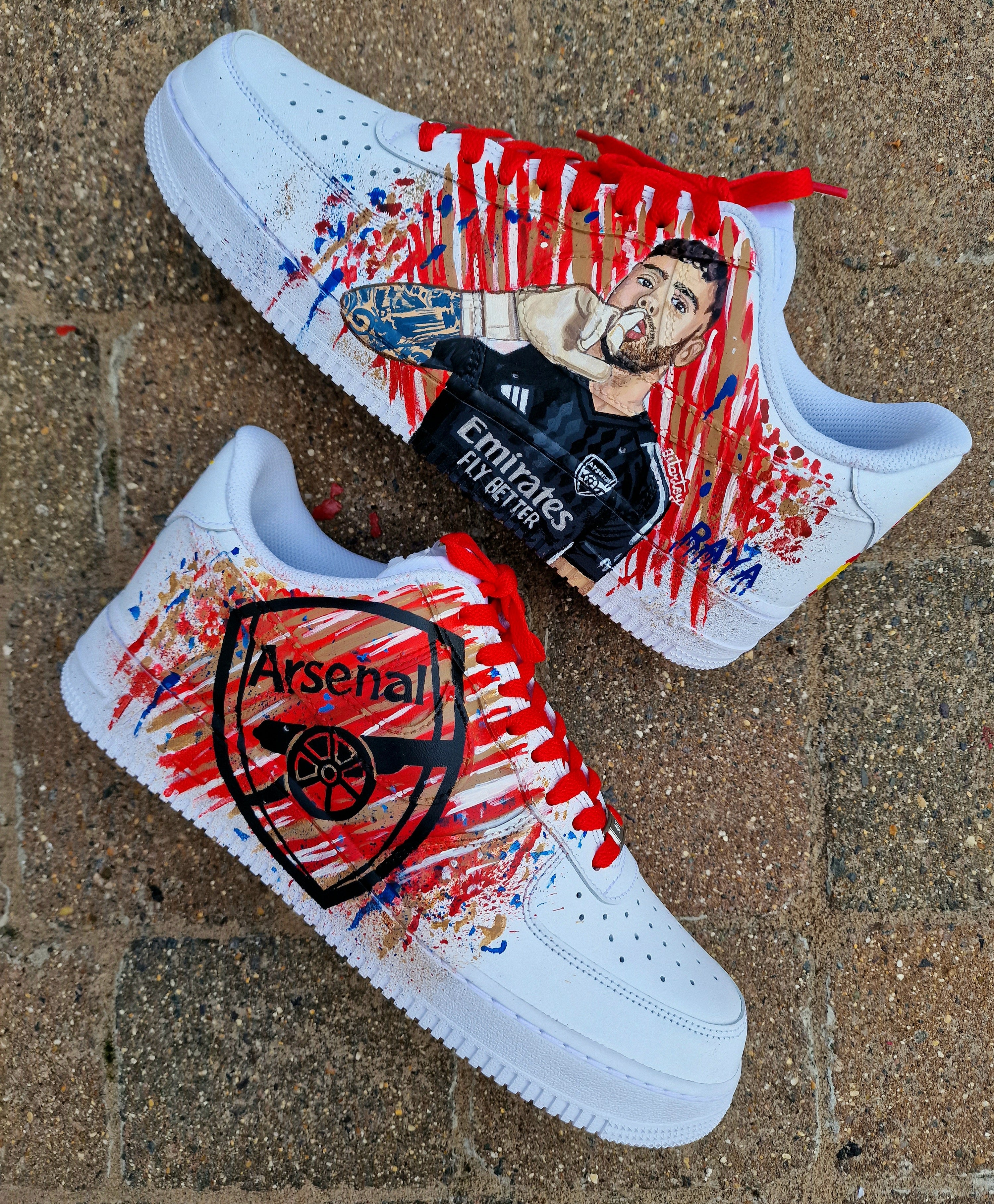 Hand-painted custom Nike Air Force 1 sneakers featuring David Raya in Arsenal goalkeeper kit, Arsenal crest, Spanish flag and colors, vibrant football-inspired splatter artwork, crafted by Gitartsy.


