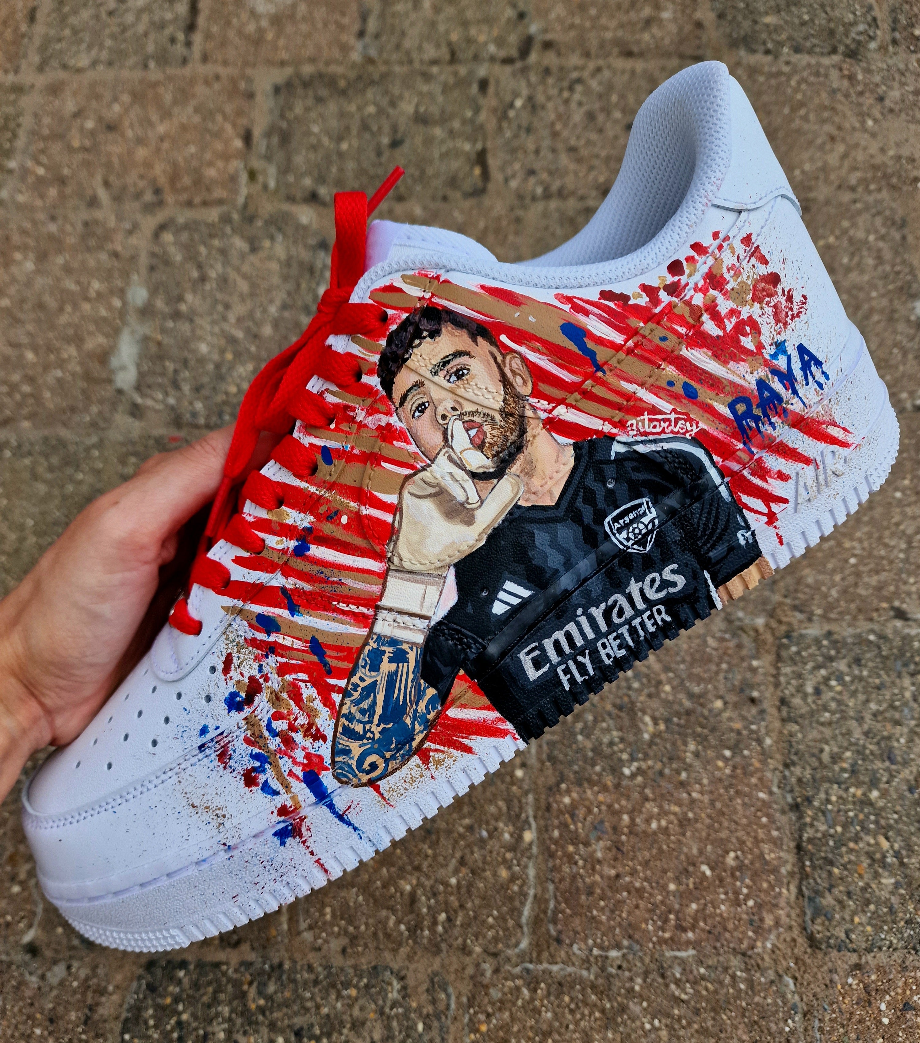 Hand-painted custom Nike Air Force 1 sneakers featuring David Raya in Arsenal goalkeeper kit, Arsenal crest, Spanish flag and colors, vibrant football-inspired splatter artwork, crafted by Gitartsy.

