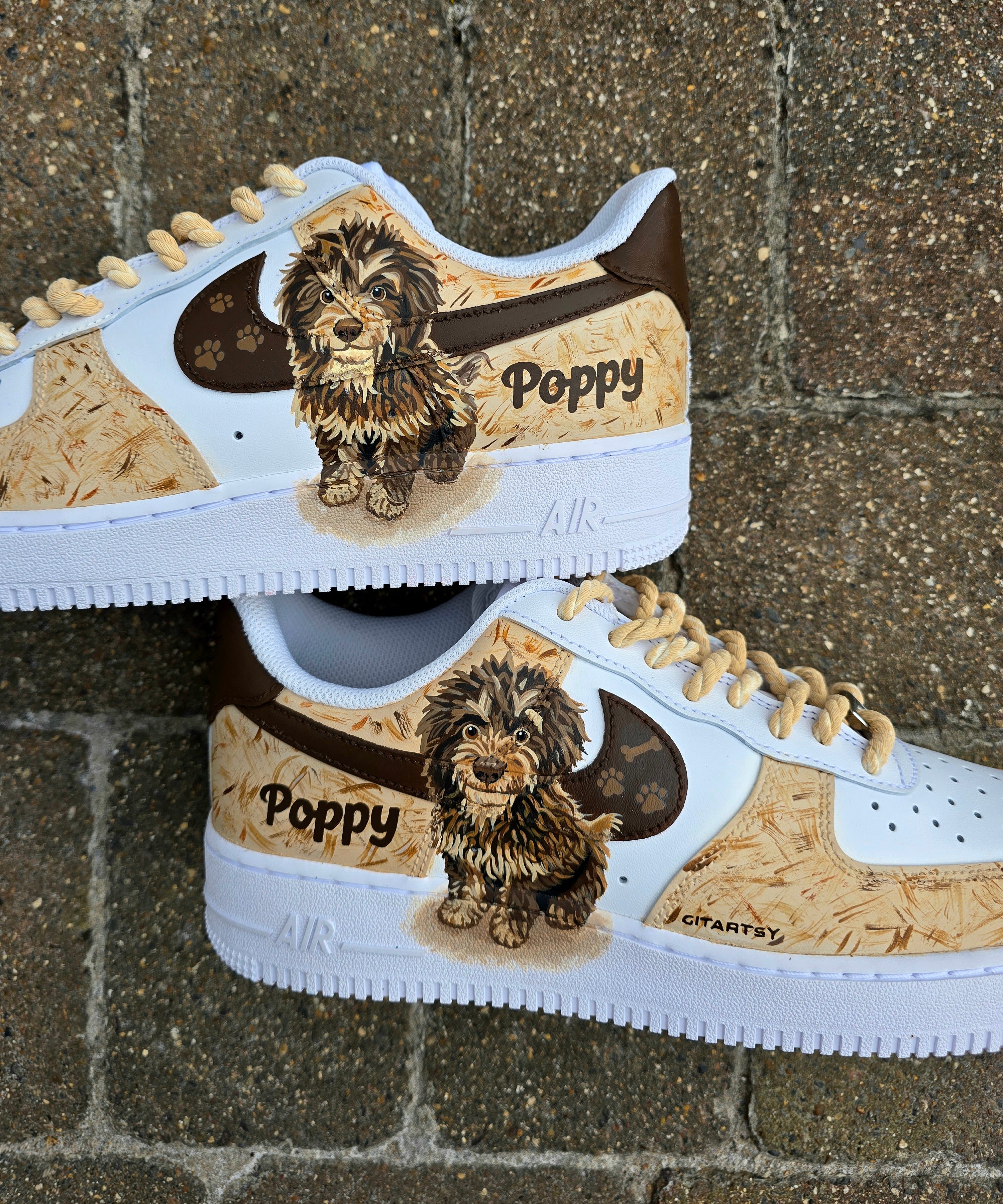 Custom hand-painted Nike Air Force 1 sneakers featuring a detailed portrait of a fluffy brown dog named Poppy 🐶. The shoes have a white leather base with light brown textured paint accents and a rich chocolate-brown swoosh adorned with paw prints 🐾. The pet’s name is written in a playful font, and tan rope laces add a rustic touch. Perfect for showcasing love for your pet in a stylish and artistic way.


