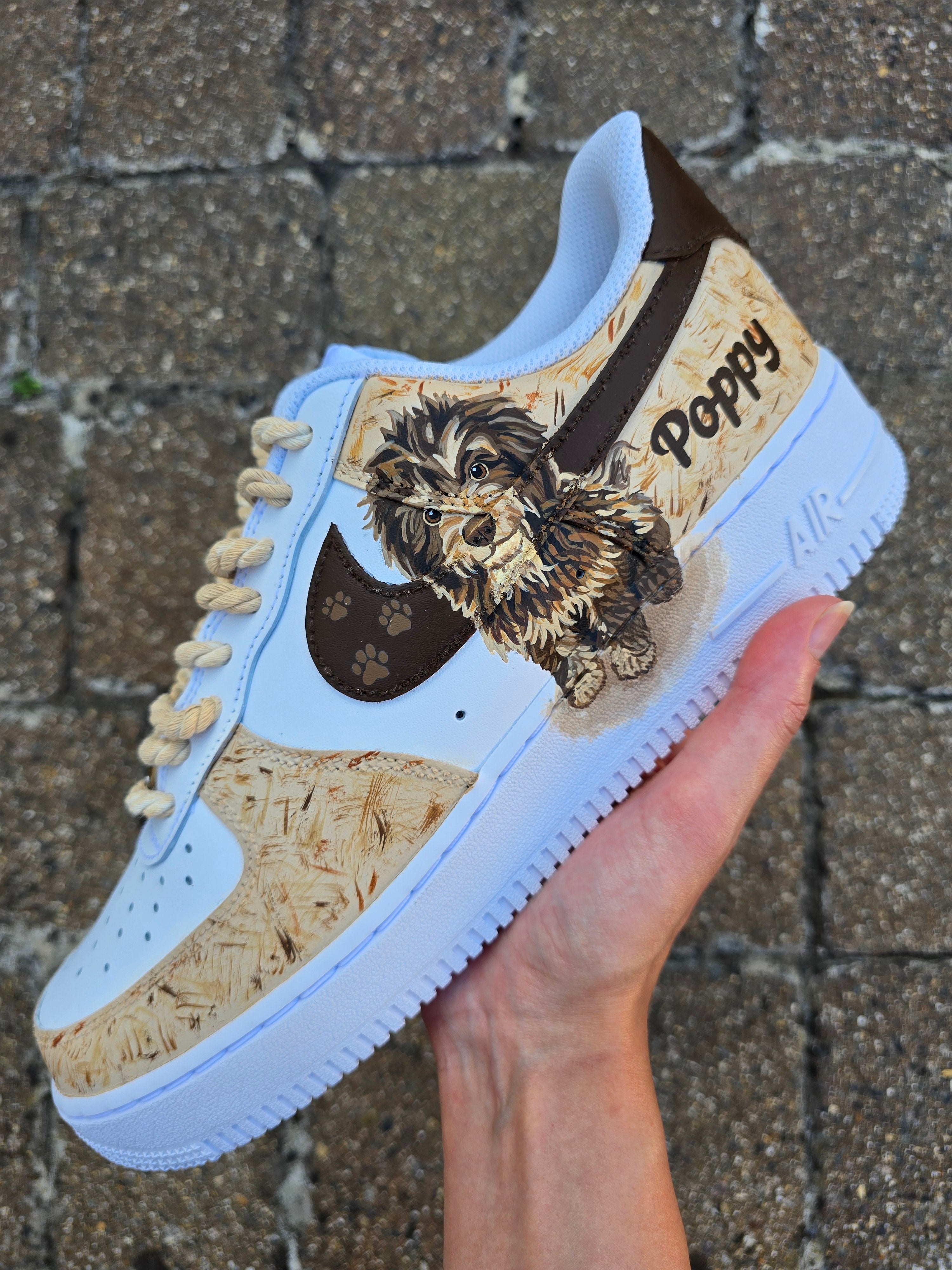 Custom hand-painted Nike Air Force 1 sneakers featuring a detailed portrait of a fluffy brown dog named Poppy 🐶. The shoes have a white leather base with light brown textured paint accents and a rich chocolate-brown swoosh adorned with paw prints 🐾. The pet’s name is written in a playful font, and tan rope laces add a rustic touch. Perfect for showcasing love for your pet in a stylish and artistic way.

