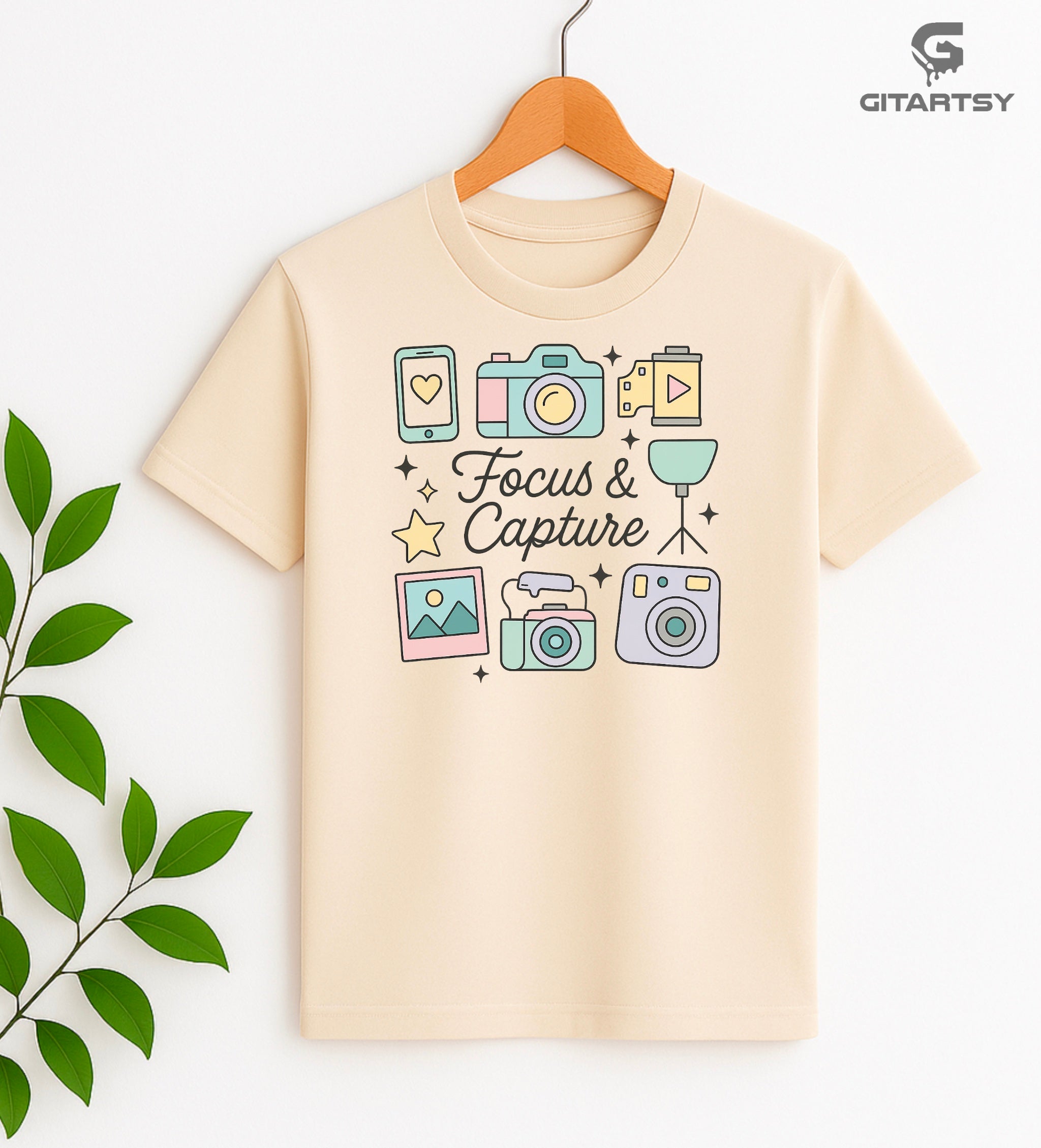 Pastel aesthetic "Focus & Capture" graphic T-shirt for photographers and videographers, featuring illustrated cameras, tripod, polaroid, smartphone, and video icons on a soft cotton tee in neutral and pastel shades.