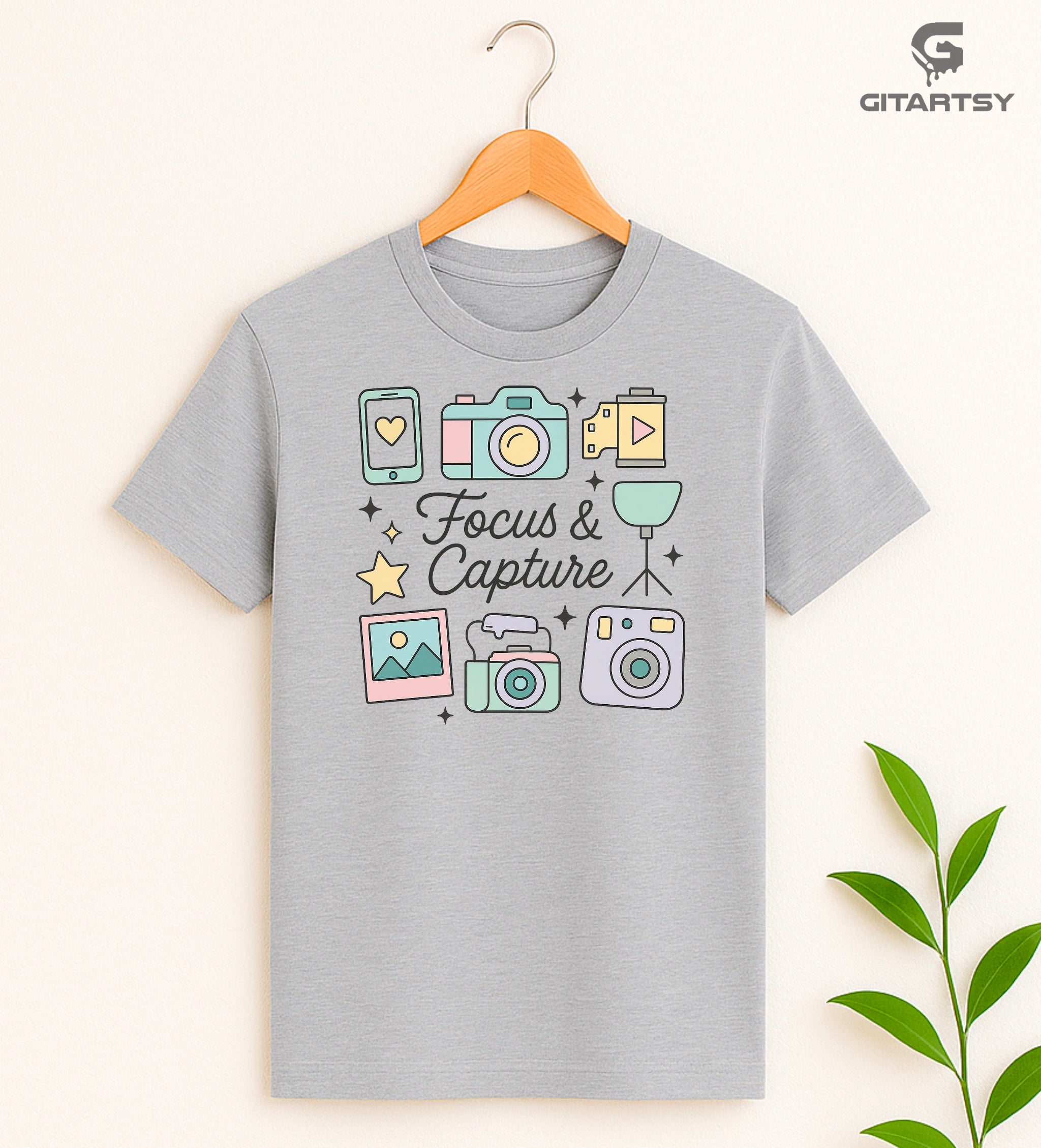 Pastel aesthetic "Focus & Capture" graphic T-shirt for photographers and videographers, featuring illustrated cameras, tripod, polaroid, smartphone, and video icons on a soft cotton tee in neutral and pastel shades.