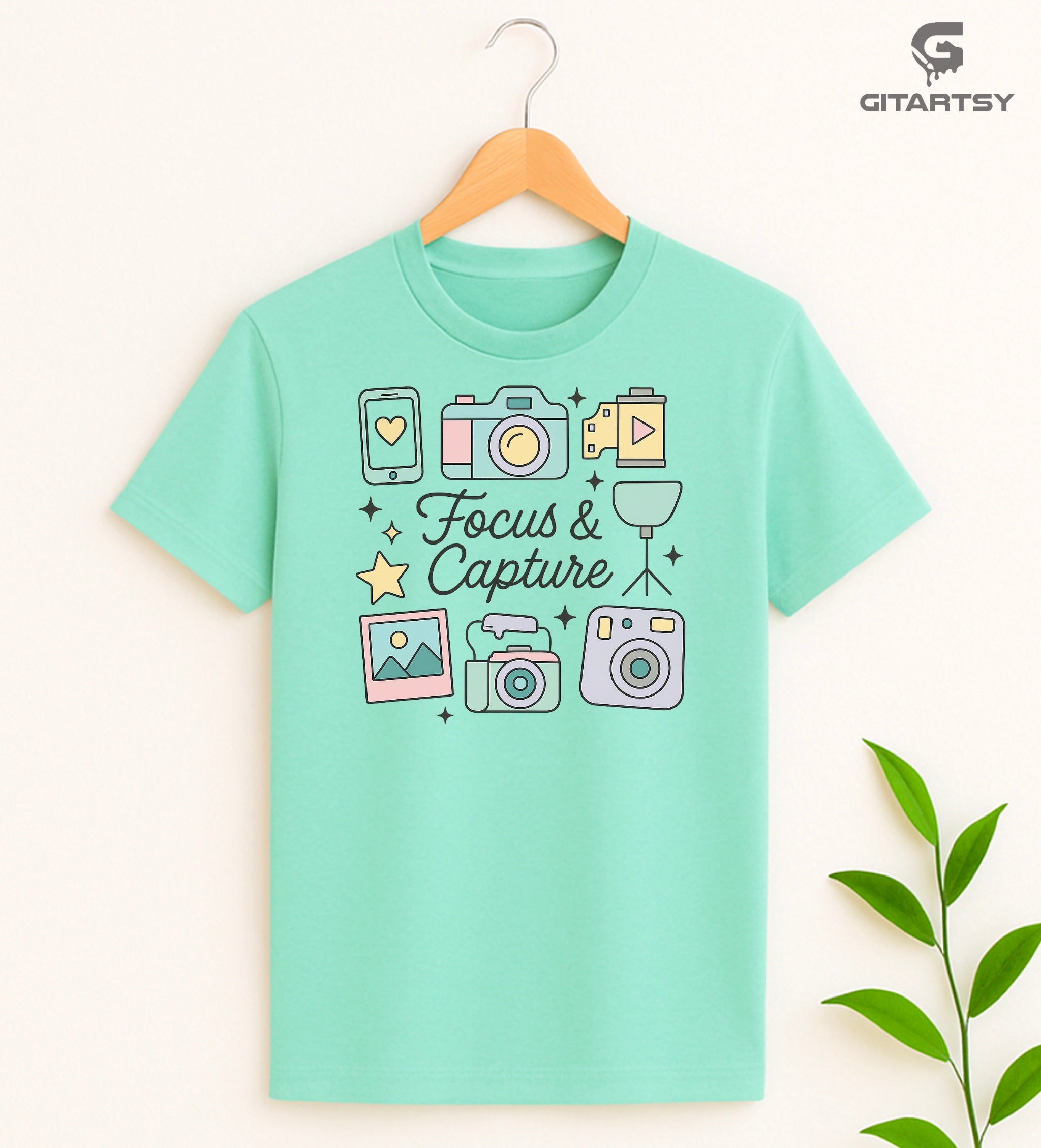 Pastel aesthetic "Focus & Capture" graphic T-shirt for photographers and videographers, featuring illustrated cameras, tripod, polaroid, smartphone, and video icons on a soft cotton tee in neutral and pastel shades.