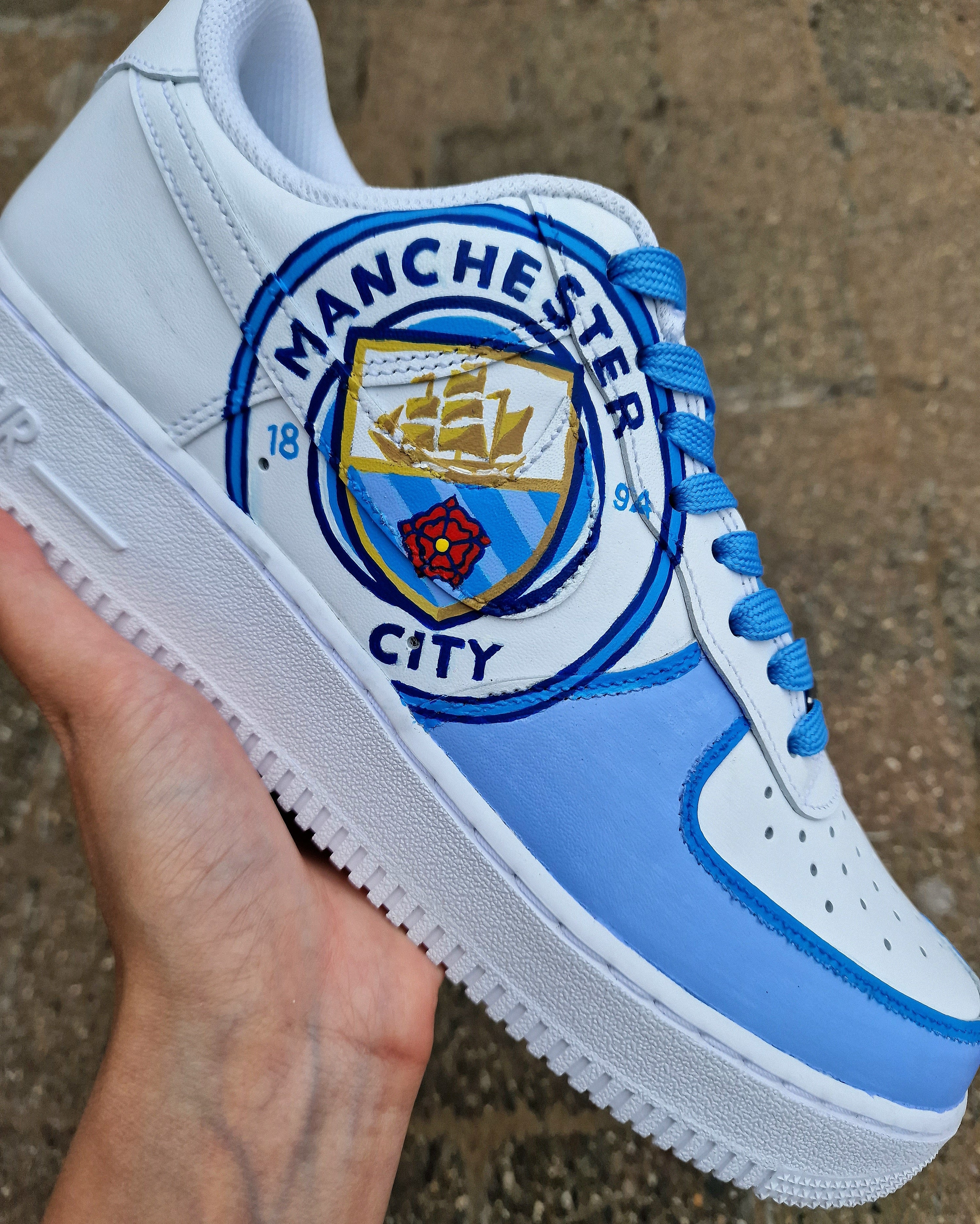 A pair of custom hand-painted Nike Air Force 1 sneakers featuring detailed artwork of the Manchester City Football Club crest, signature blue hues, striped swoosh, and personalized finishing, crafted by Gitartsy for football fans and sneaker collectors.

