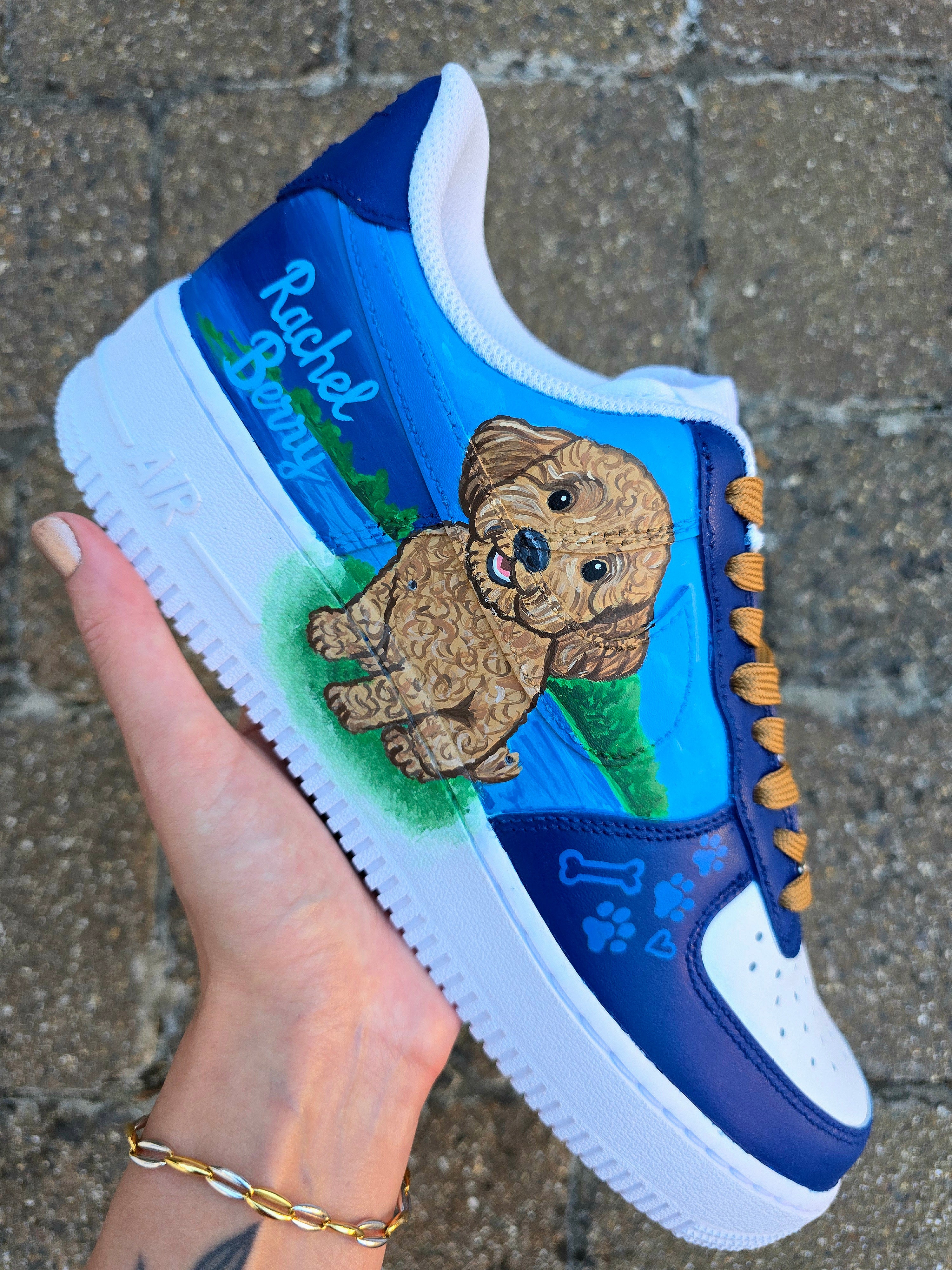 Custom Nike Air Force 1 sneakers 🐾 hand-painted with a detailed toy poodle pet portrait, featuring a caramel-colored curly-haired dog sitting in a grassy field with a blue sky and lake background. Includes the pet’s name in stylish script, paw prints, bones, and heart icons, all painted on a blue and navy color-block base with golden-brown laces. Each pair is personalized to match your pet’s likeness and crafted using durable, waterproof leather paints, making them wearable art for dog lovers.

